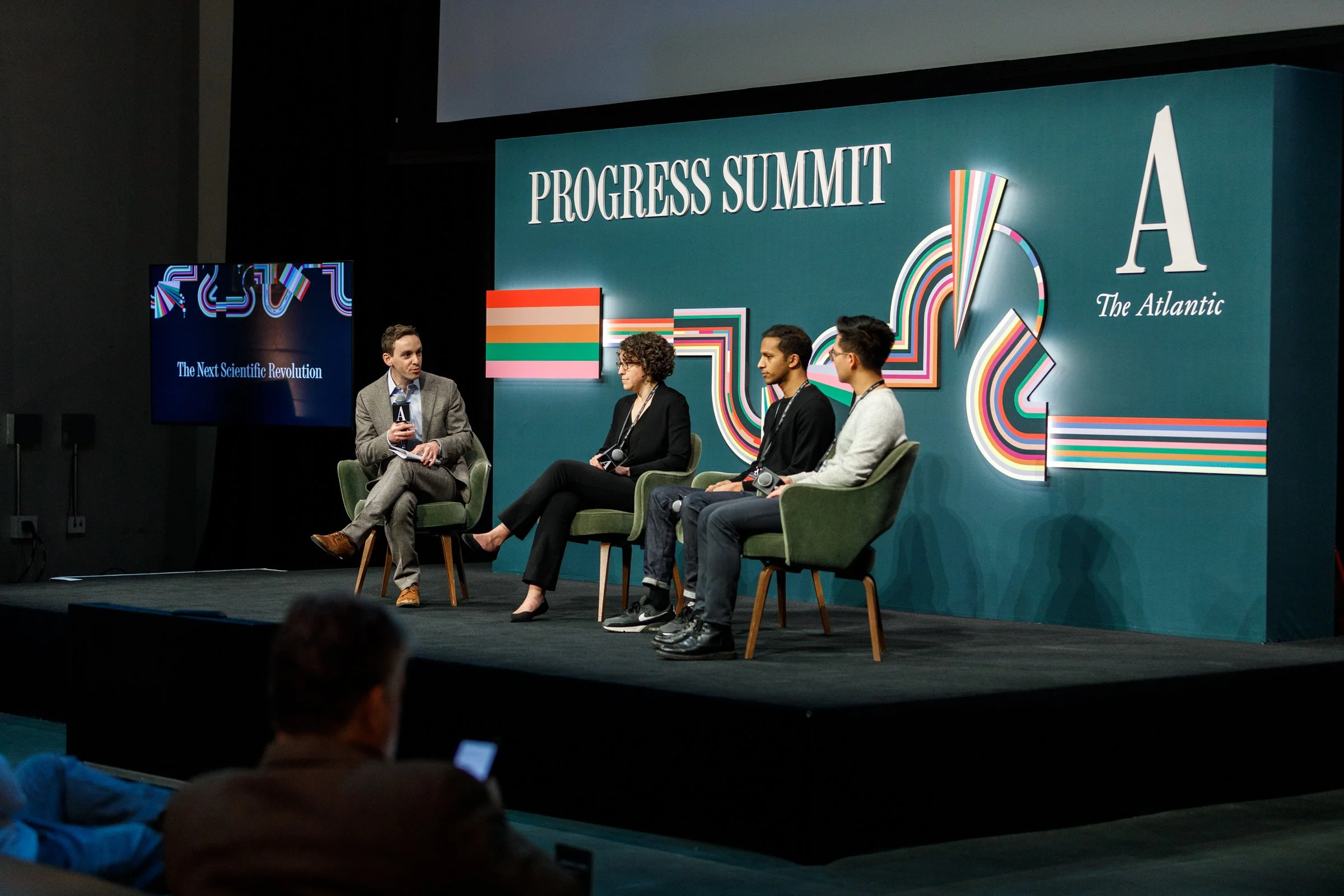 Panel discussion at Progress Summit with four speakers sitting on stage, one holding a microphone; backdrop reads "Progress Summit" and "The Atlantic"; large screen displays "The Next Scientific Revolution."
