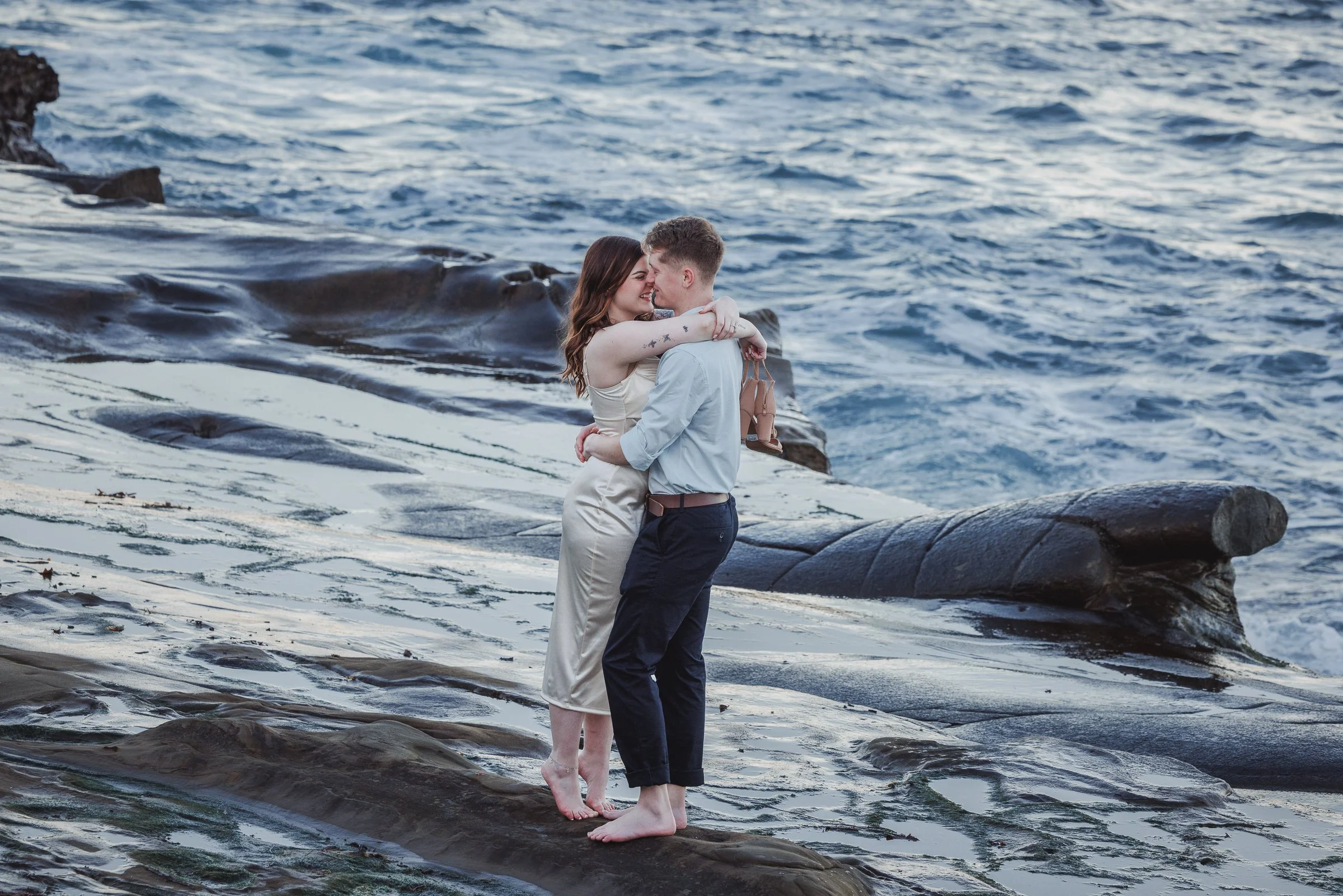 A couple embracing on a rocky shoreline by the ocean, smiling at each other.