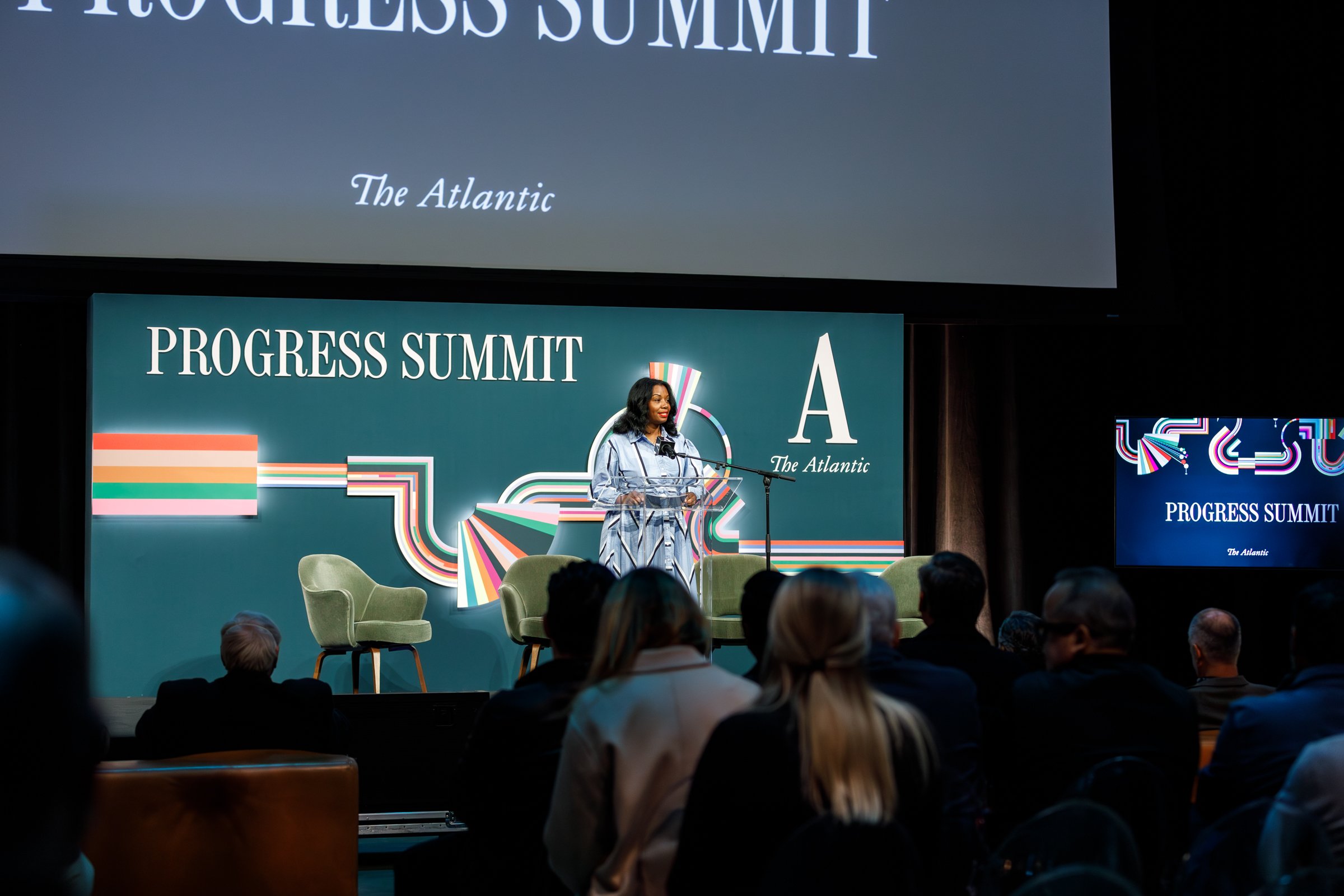 A woman speaking at a podium on stage during the Progress Summit, with a large backdrop featuring the words 'Progress Summit' and 'The Atlantic' behind her. Several audience members are seated and listening.