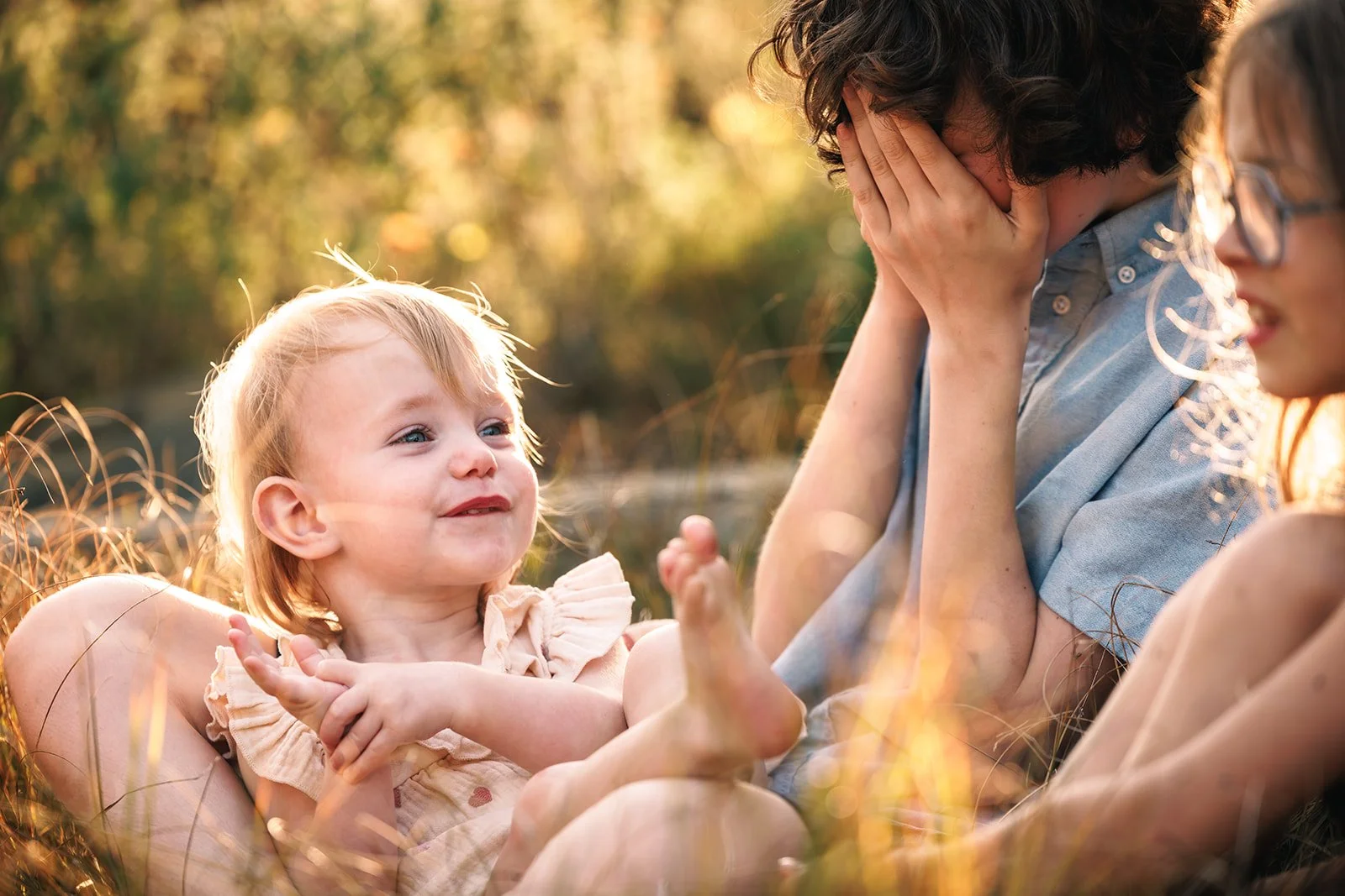 A young girl with blonde hair smiling and playing with an adult, who has curly hair and covers their face with hands, while another woman with glasses watches nearby in a natural outdoor setting during sunset.