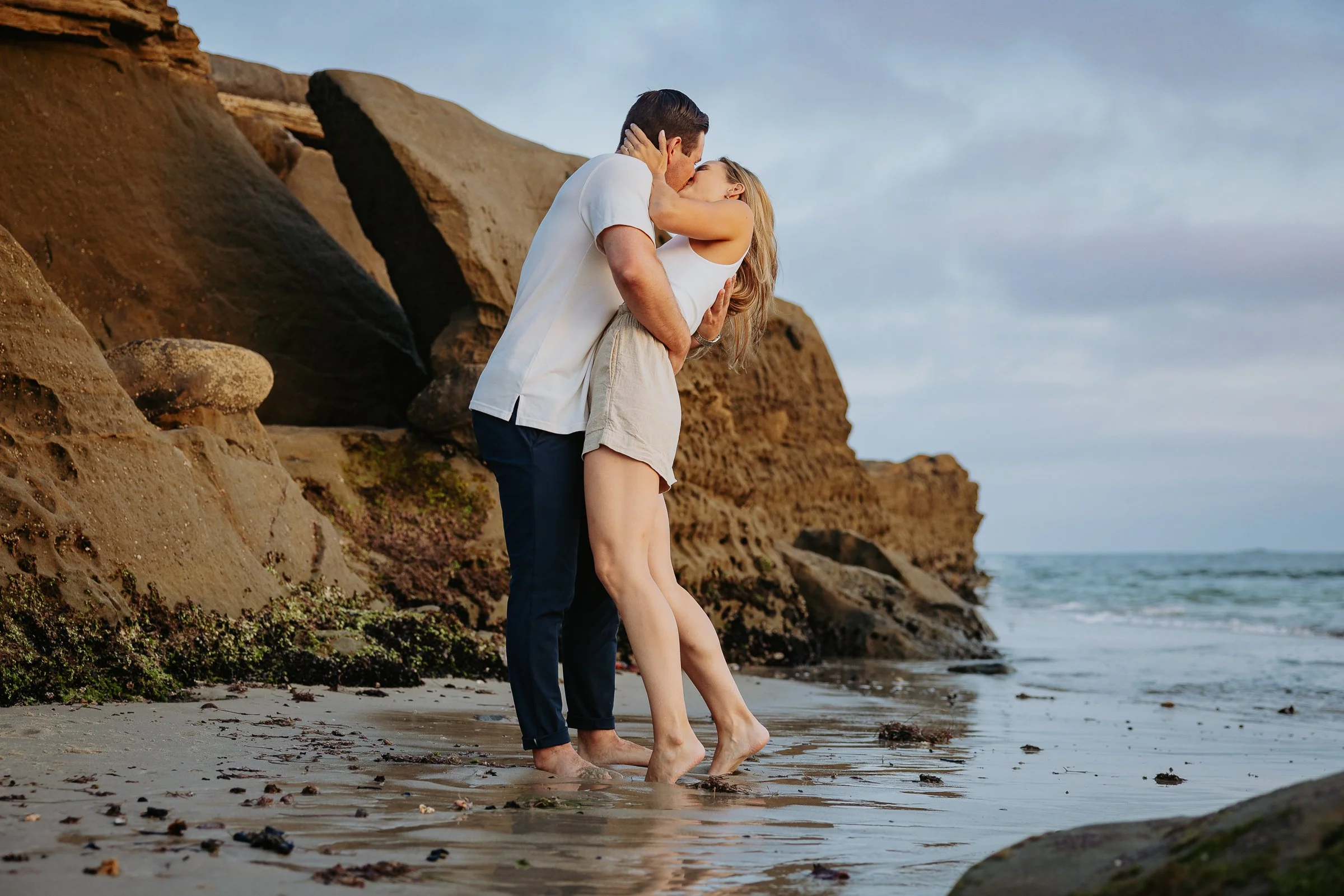A couple kissing on a beach near rocky cliffs, with ocean waves in the background.