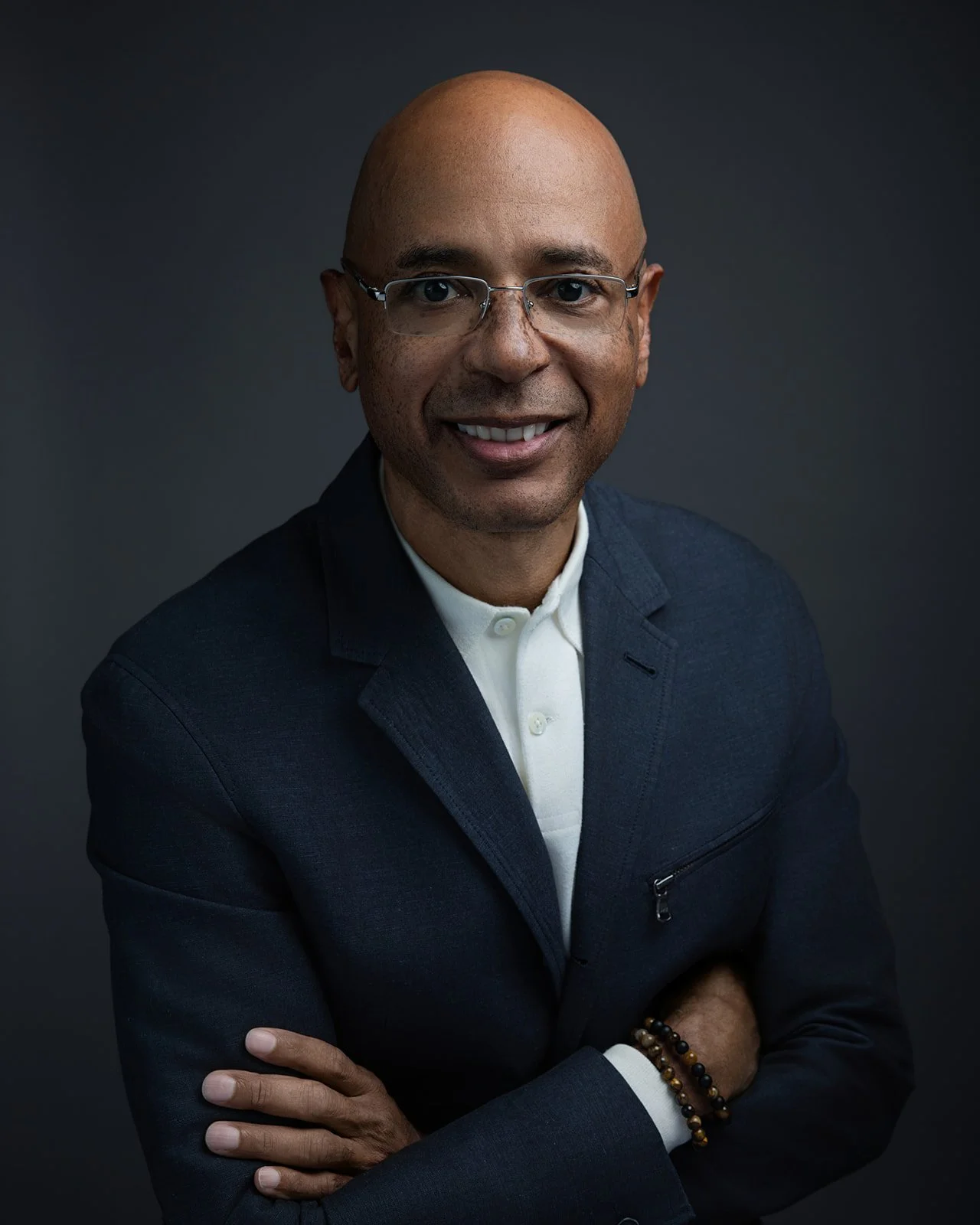 A professional man with glasses, wearing a dark blazer over a white shirt, smiling, with arms crossed, against a dark background.