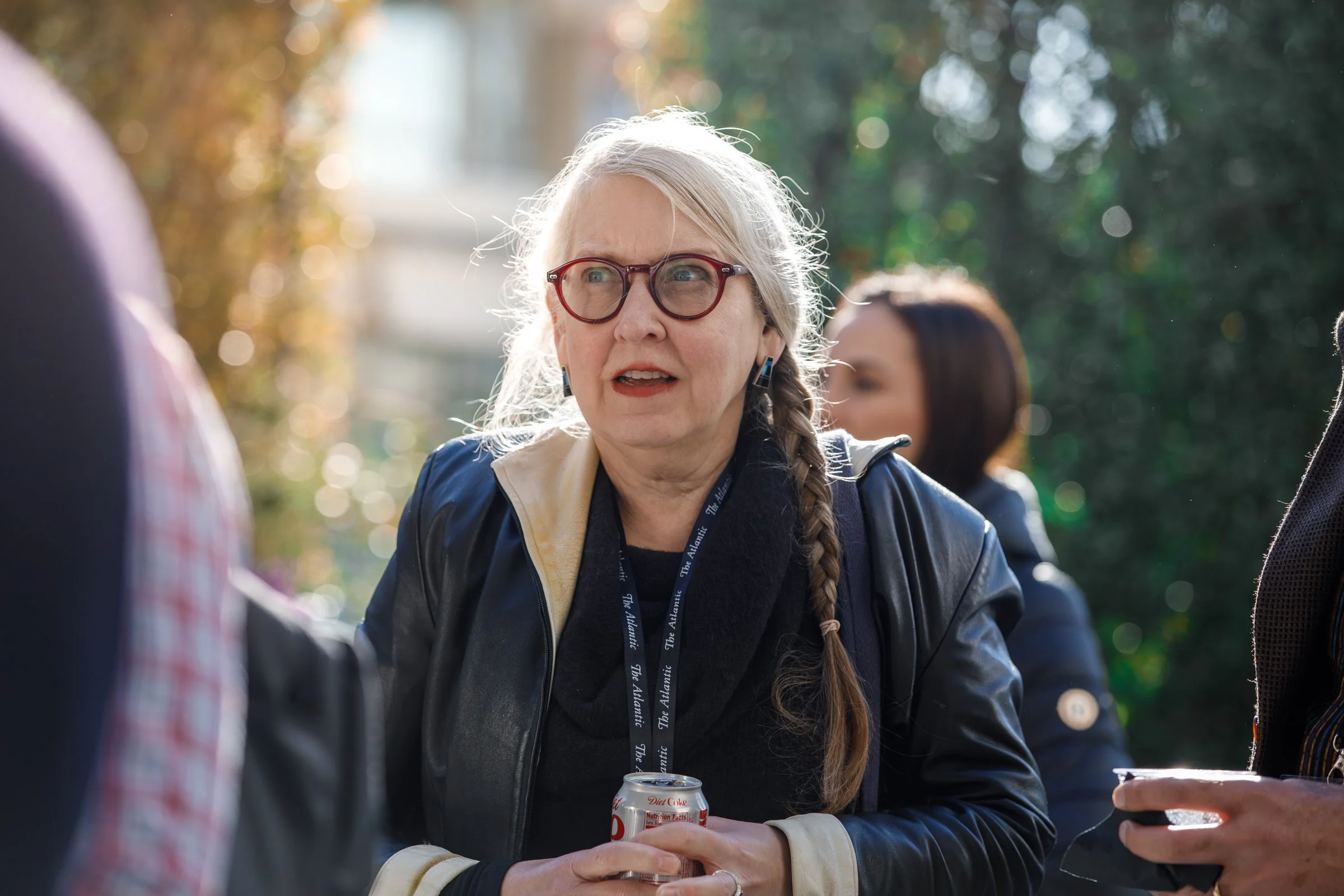 An elderly woman with gray hair, glasses, and a braid talking outdoors, holding a soda can, with other people nearby and trees in the background.