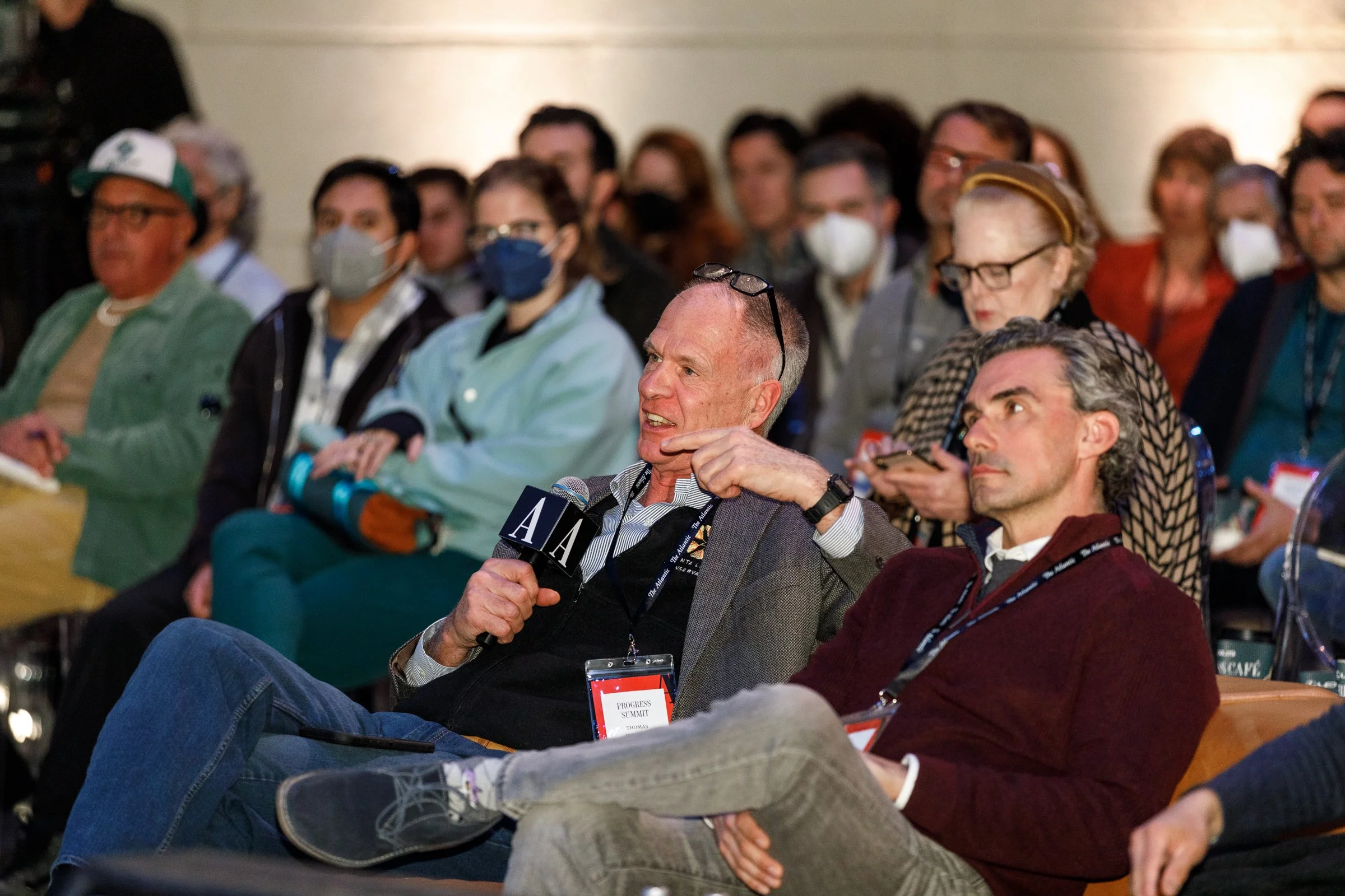 A man holding a microphone labeled with the letter 'A' is speaking at a conference, surrounded by an audience of masked and unmasked attendees listening attentively.
