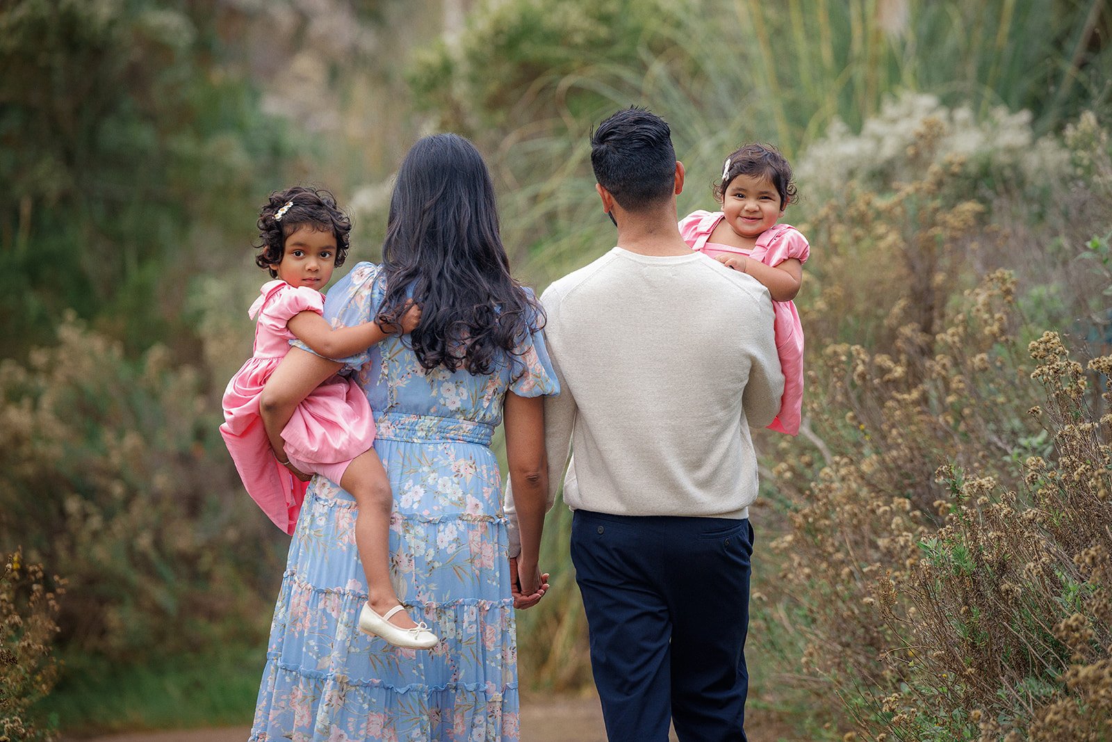 Family walking outdoors on a trail, with two girls in pink dresses being carried by their parents.