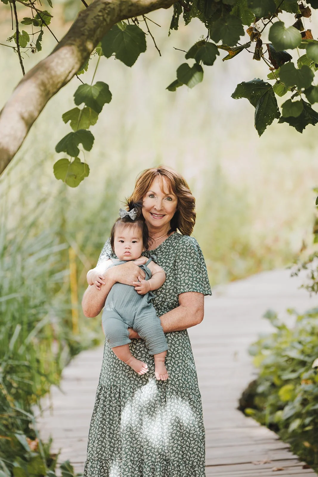 A smiling woman holding a young child with a bow in her hair, standing on a wooden path surrounded by greenery.