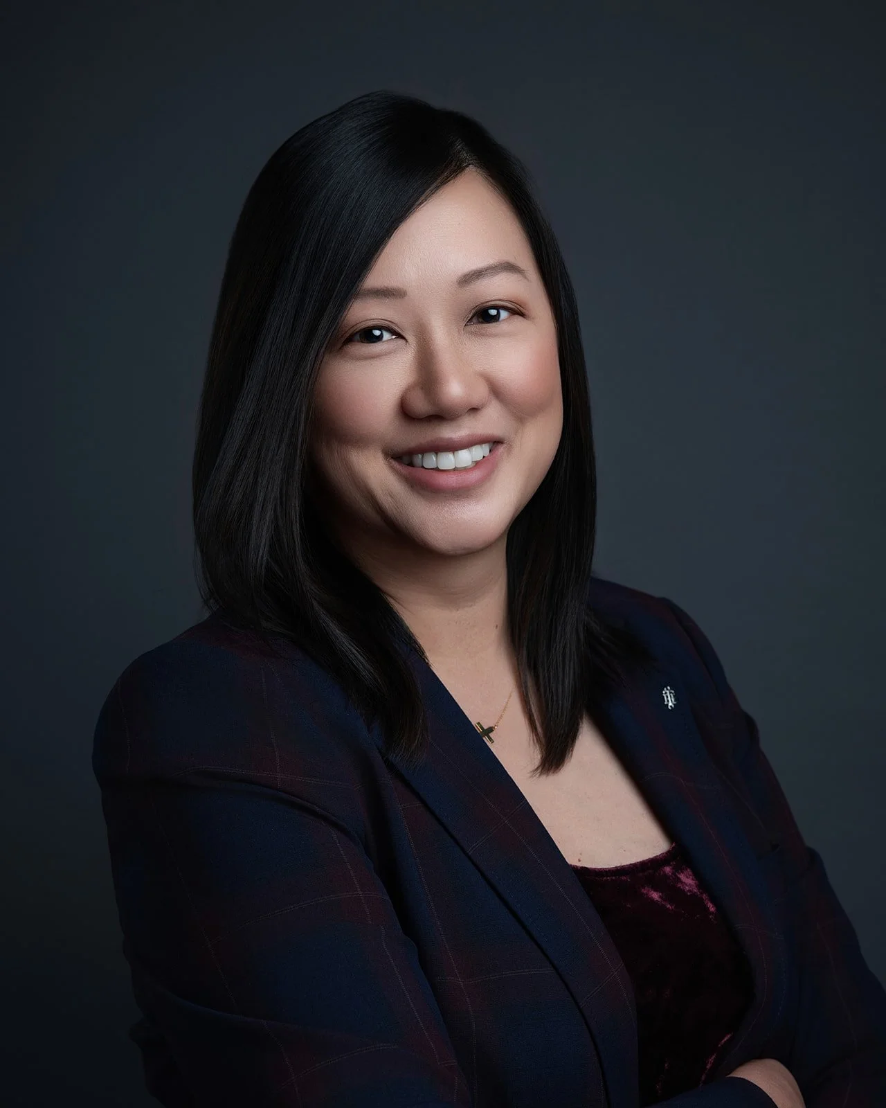 Professional portrait of an Asian woman with shoulder-length black hair, smiling, wearing a dark blazer and top, against a dark background.