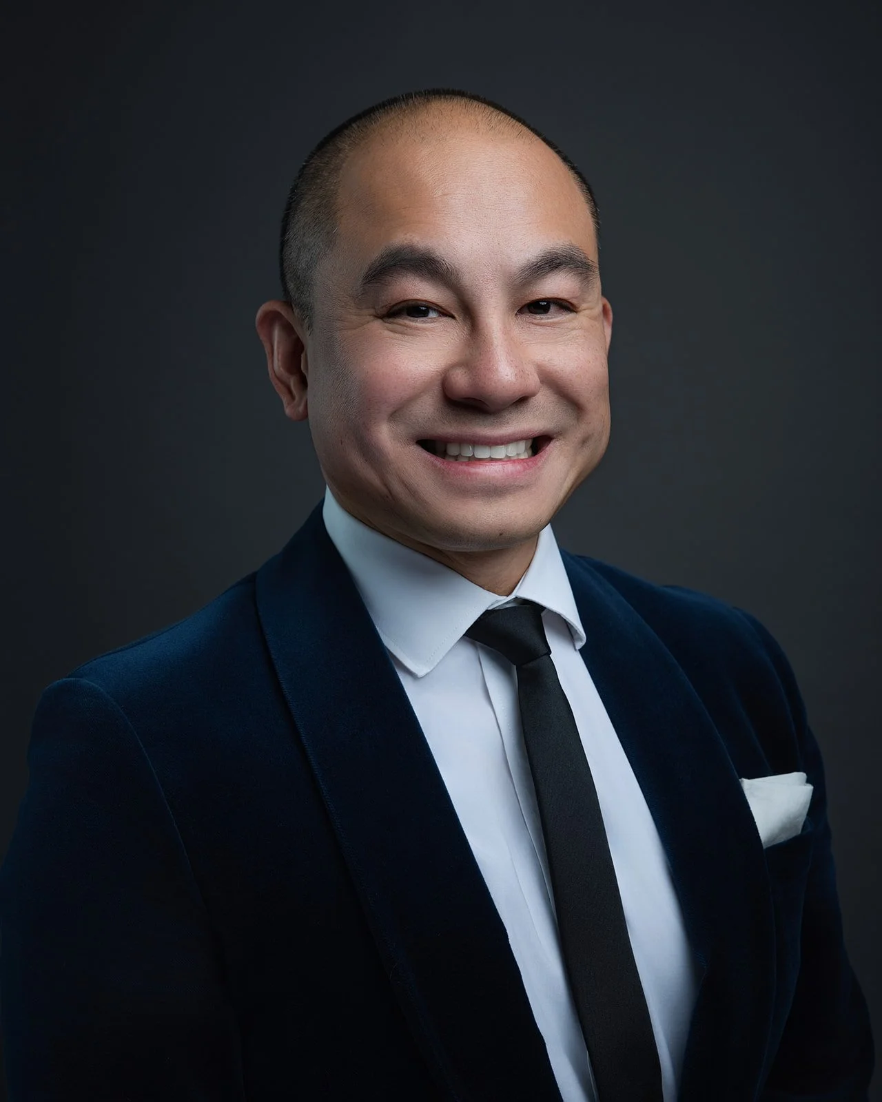 Professional portrait of an Asian man with a shaved head, wearing a dark blue velvet blazer, white shirt, and black tie, smiling against a dark background.