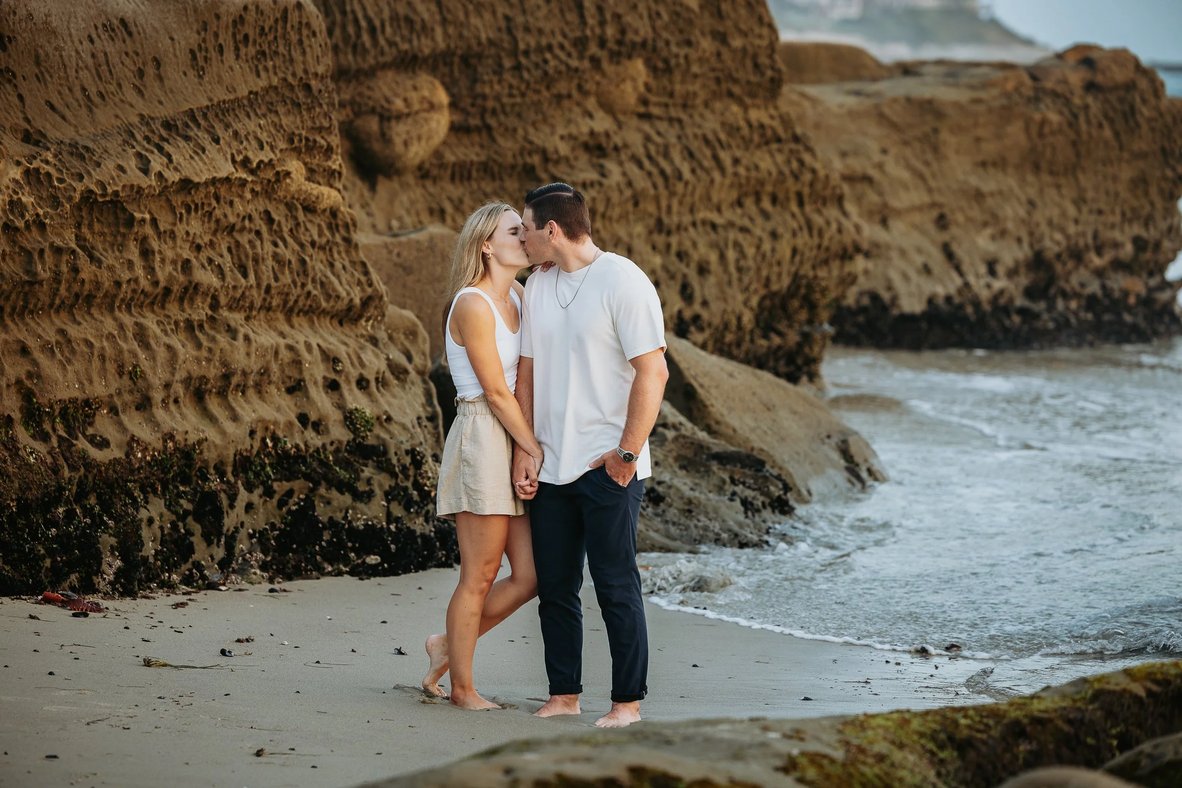 A couple kisses on the beach near large rock formations.