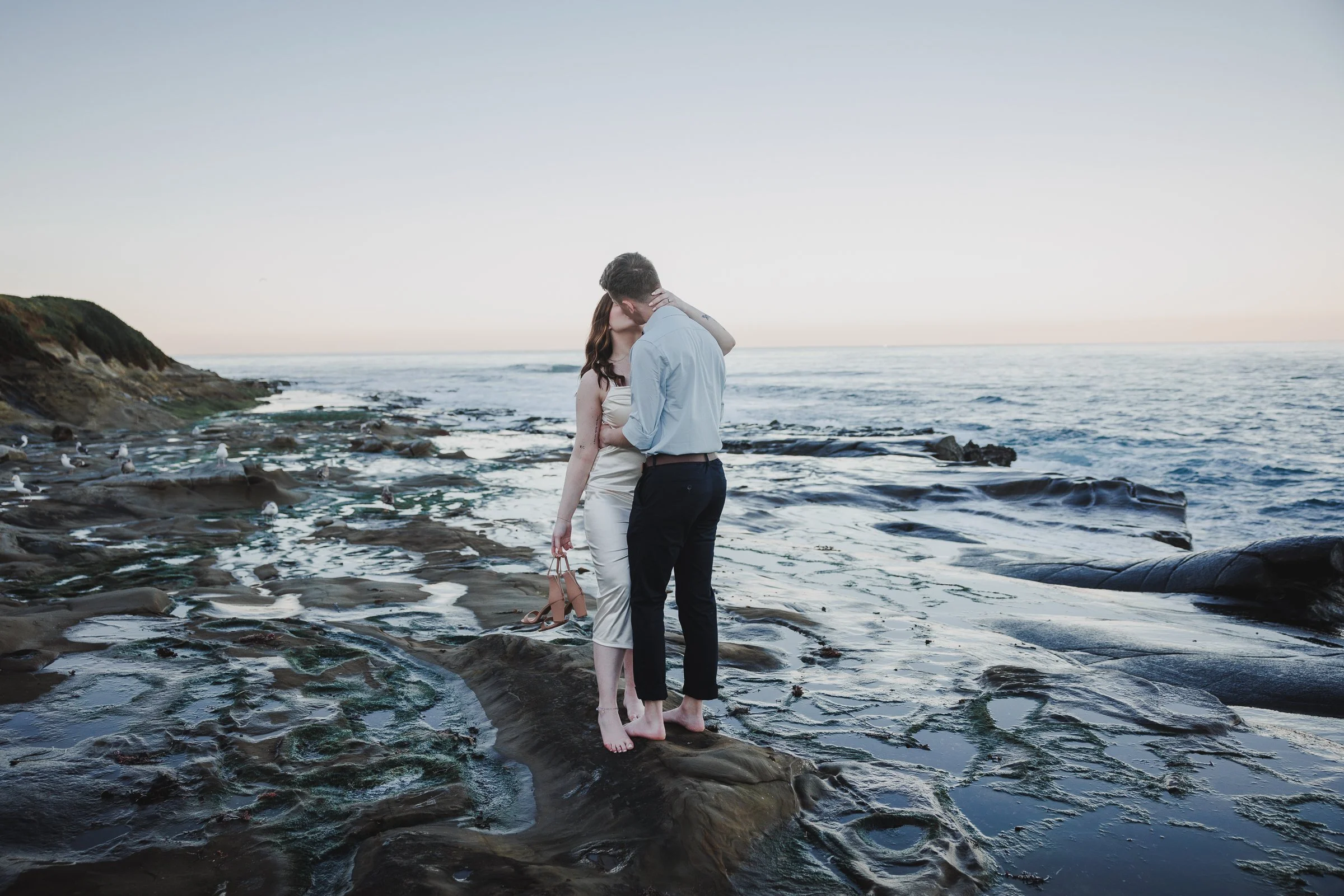 A couple is kissing on a rocky beach with the ocean in the background. The woman is wearing a light-colored dress, holding shoes in her hand, and the man is dressed in a light shirt and dark pants.