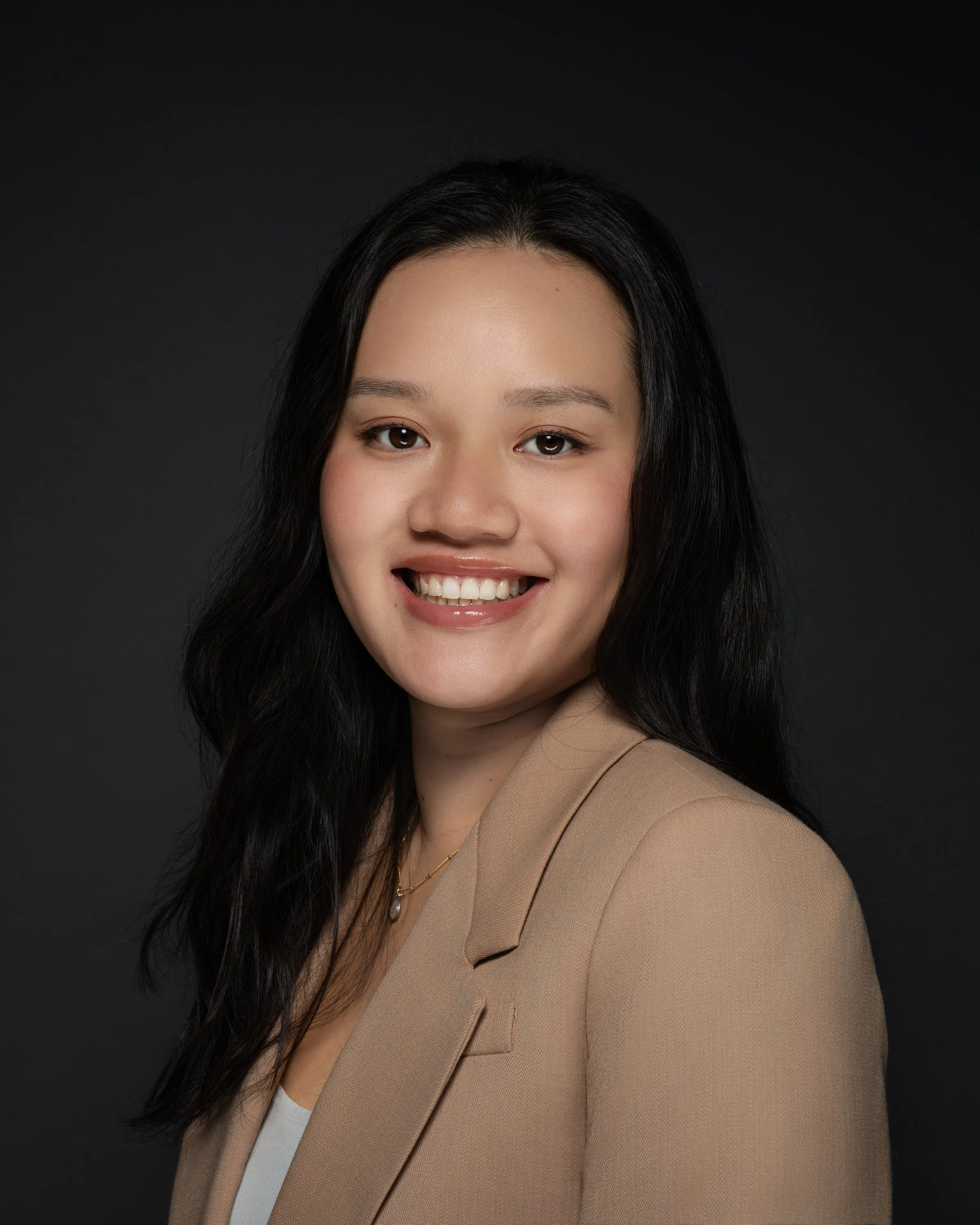 Professional headshot of a woman with long black hair smiling, wearing a beige blazer and white top, against a dark background.