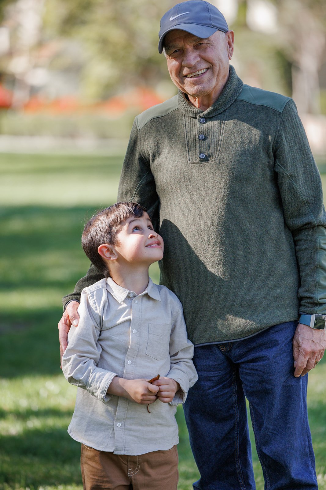 An elderly man in a green sweater and blue cap smiling while standing outdoors with a young boy in a beige shirt, looking up at him and smiling, on a grassy area with trees in the background.
