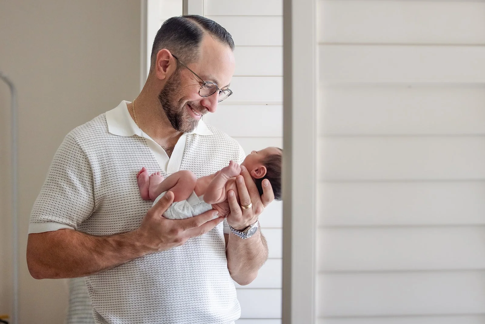 A man holding a newborn baby and smiling at it indoors with white walls.