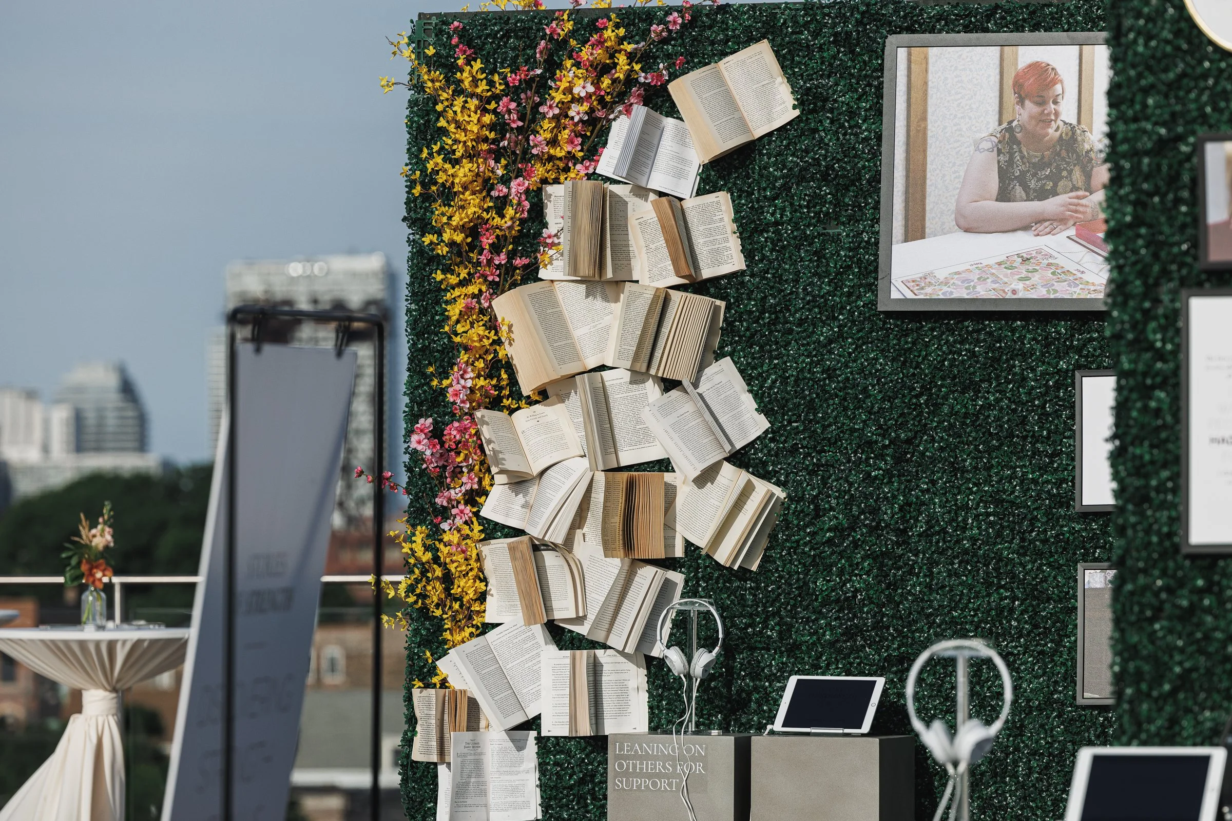 A decorative outdoor display with a green hedge wall decorated with open books, pink and yellow flowers, framed photographs, and digital devices, set against a city skyline background.