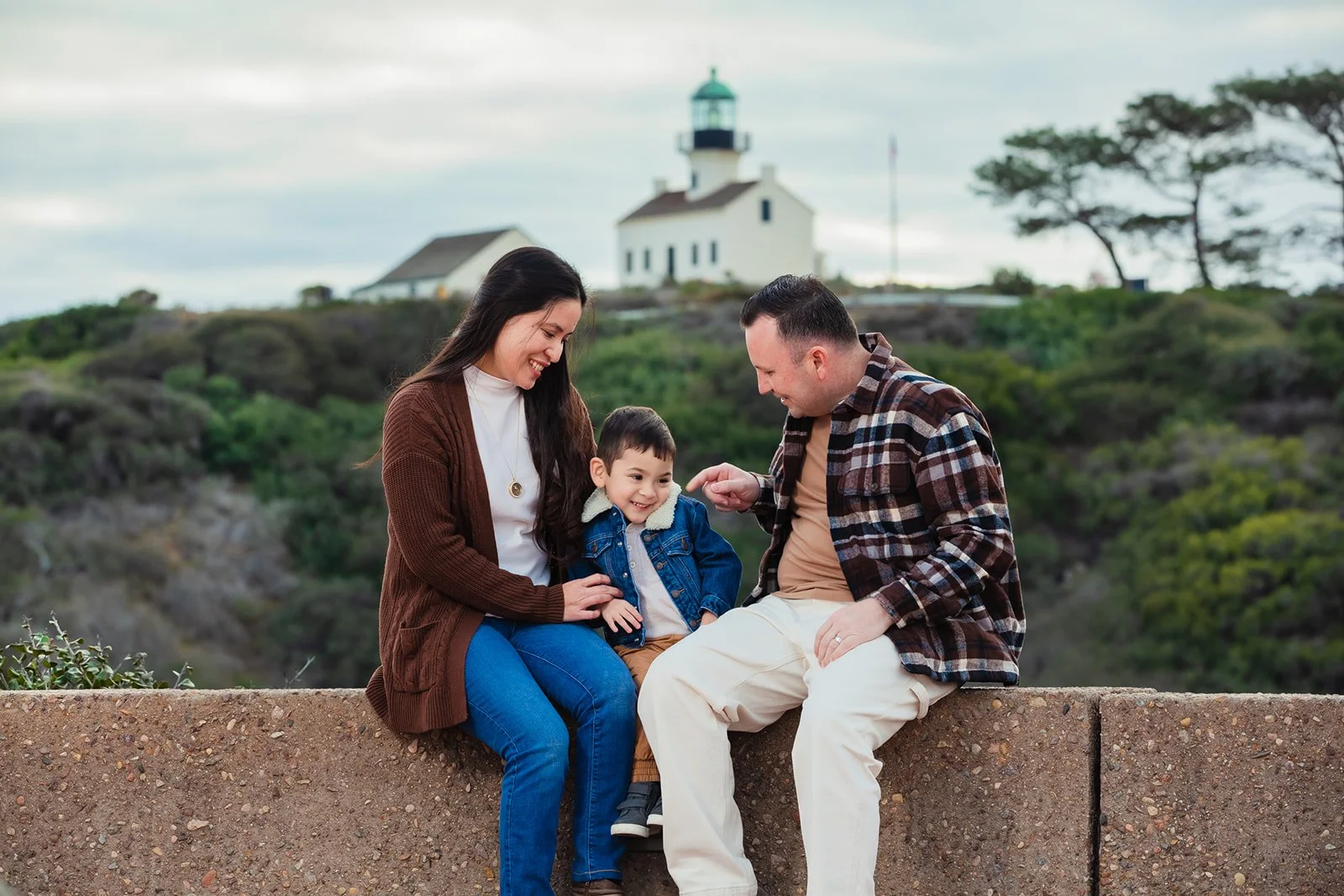 A family of three sitting on a concrete wall outdoors, with a lighthouse and green hills in the background. The woman and man are smiling at a young boy, who is sitting on the wall between them.