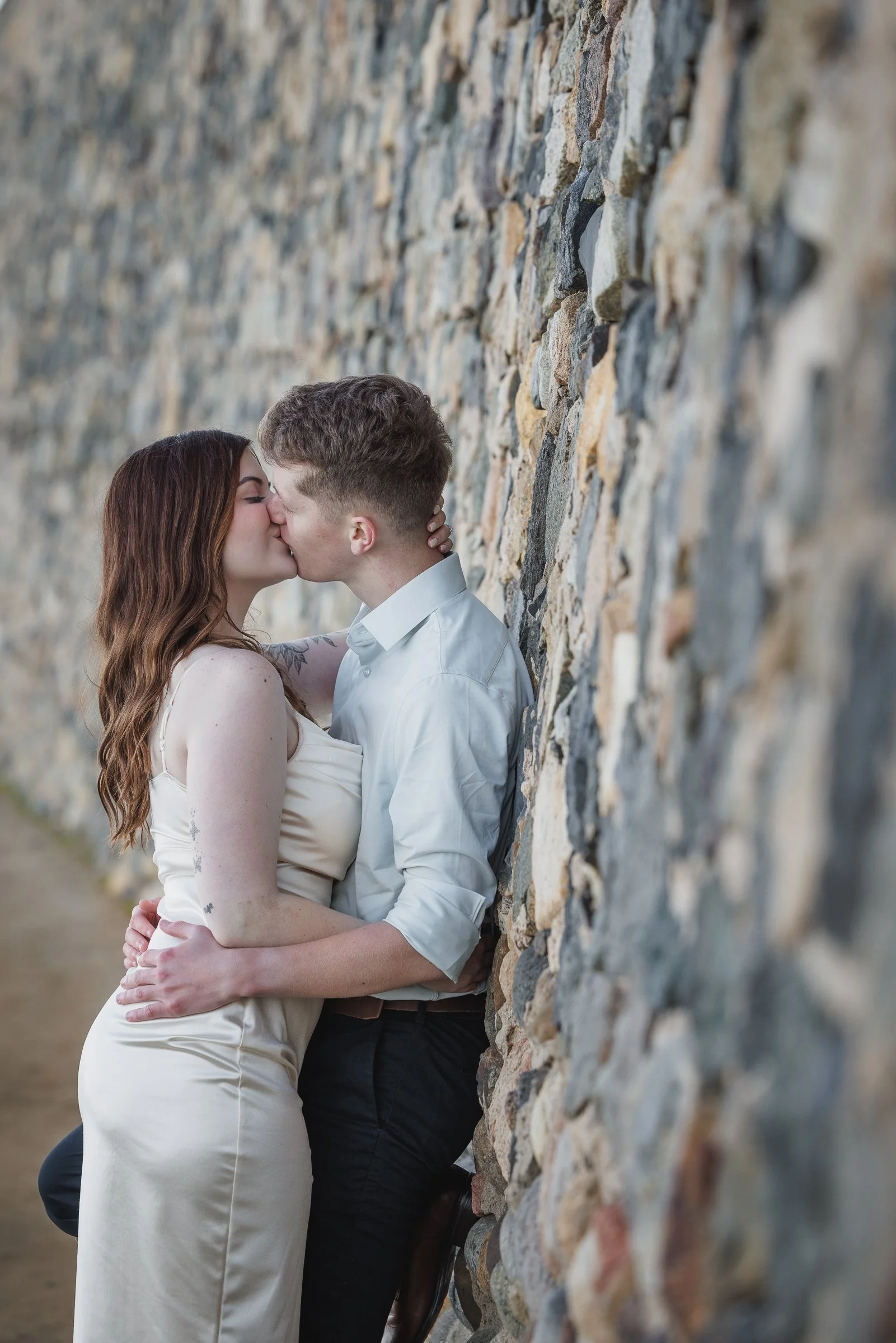 A couple kissing while standing against a stone wall, with the woman wearing a white dress and the man in a white shirt and dark pants.