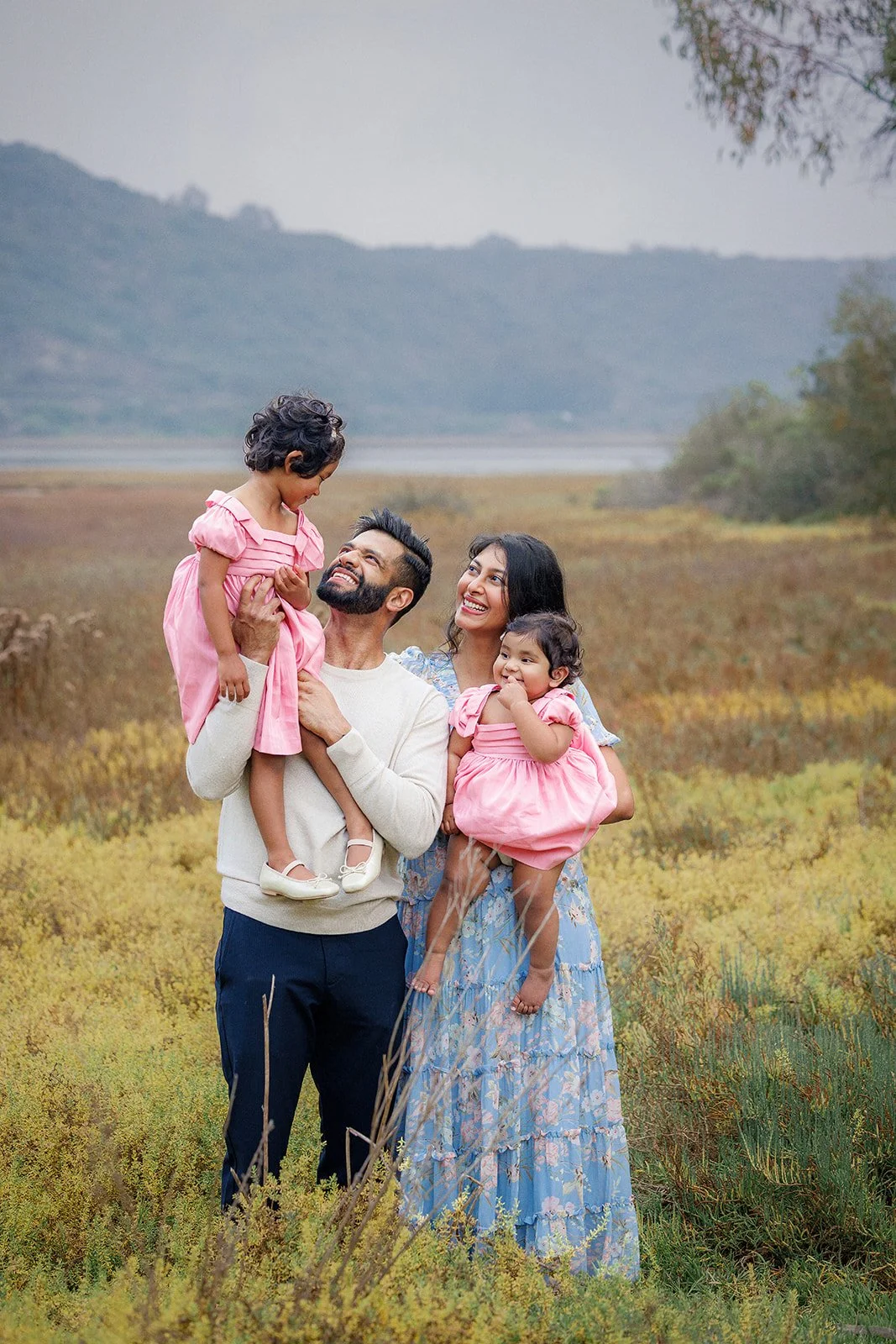 A family of four, including a father, mother, and two young daughters, standing outdoors in a field with mountains in the background. The father is holding one daughter while the mother holds the other. All are smiling and wearing casual clothing, wi