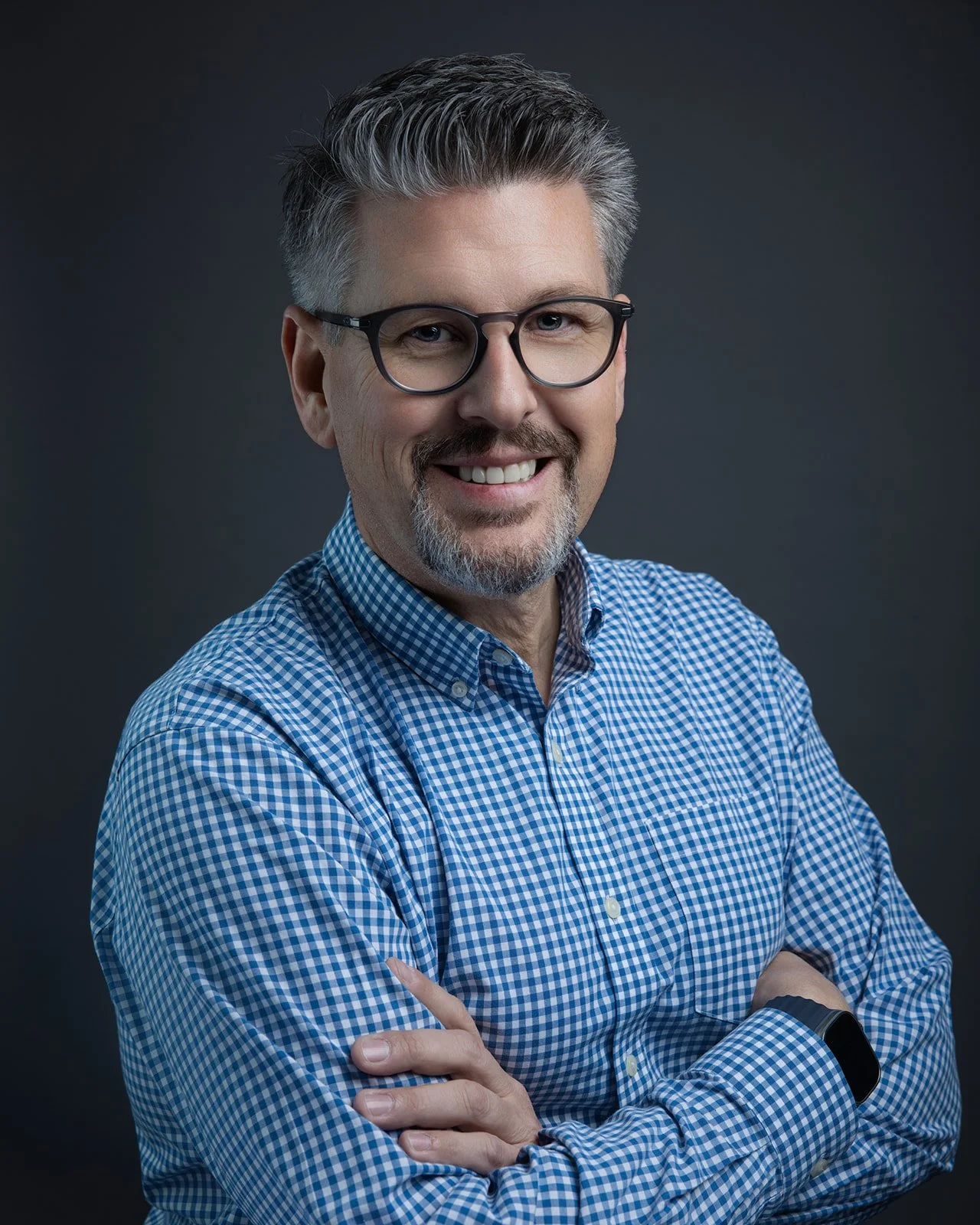 A middle-aged man with gray hair and glasses, smiling, wearing a blue and white checkered shirt, with arms crossed, standing against a dark gray background.