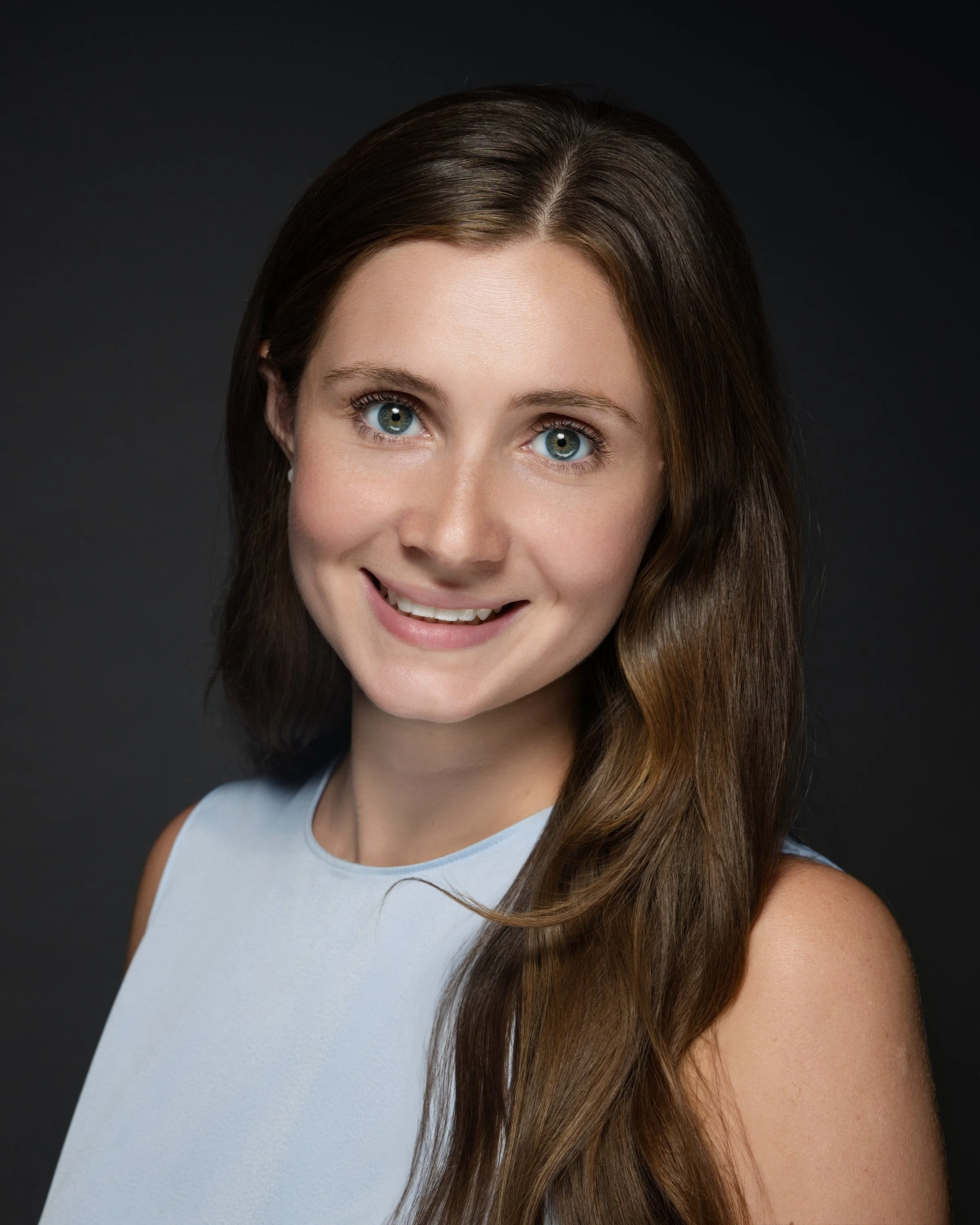 Portrait of a smiling young woman with long brown hair, blue eyes, wearing a light blue sleeveless top, against a dark background.