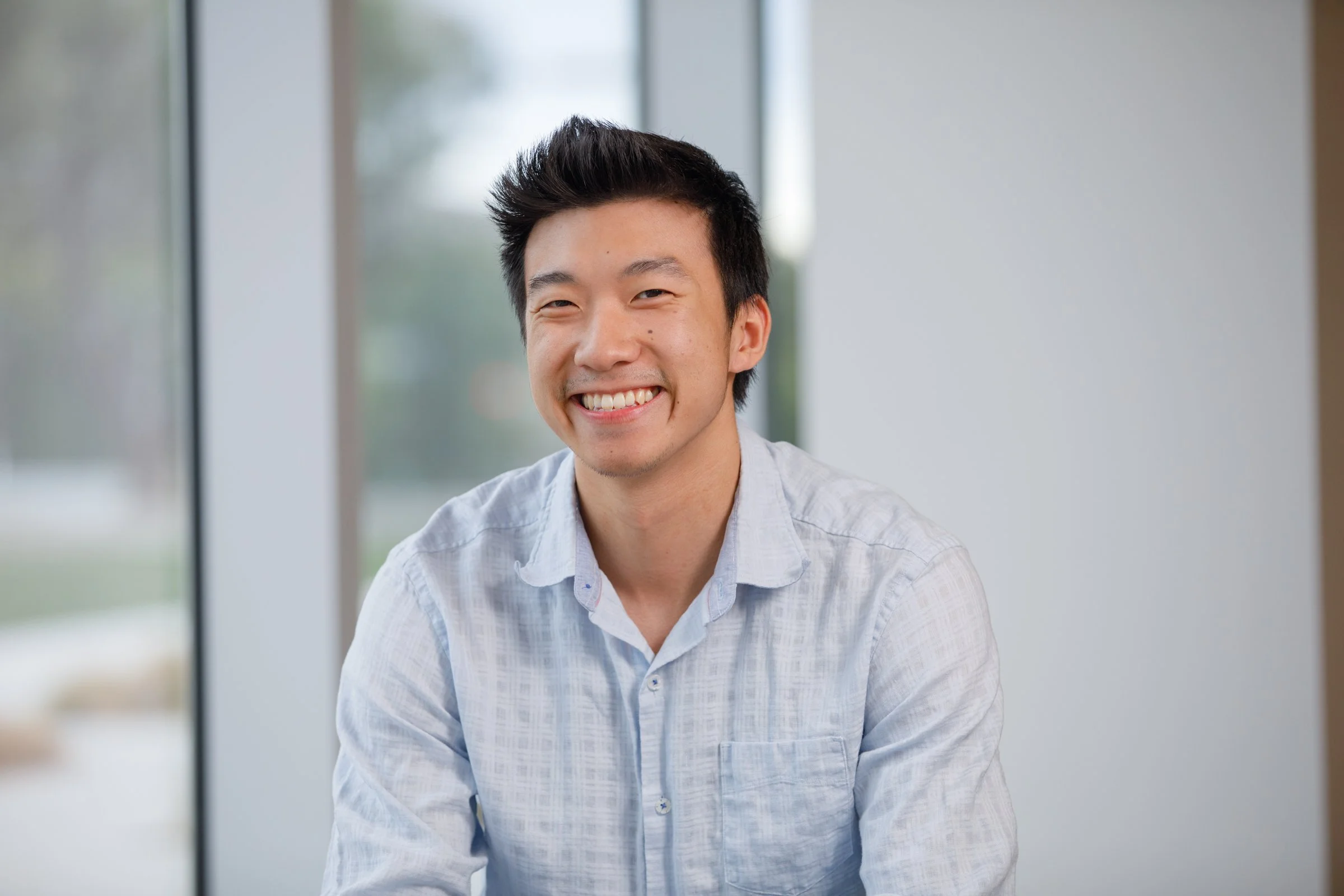 A young man with short black hair, smiling, wearing a light-colored button-up shirt, sitting indoors near a window.
