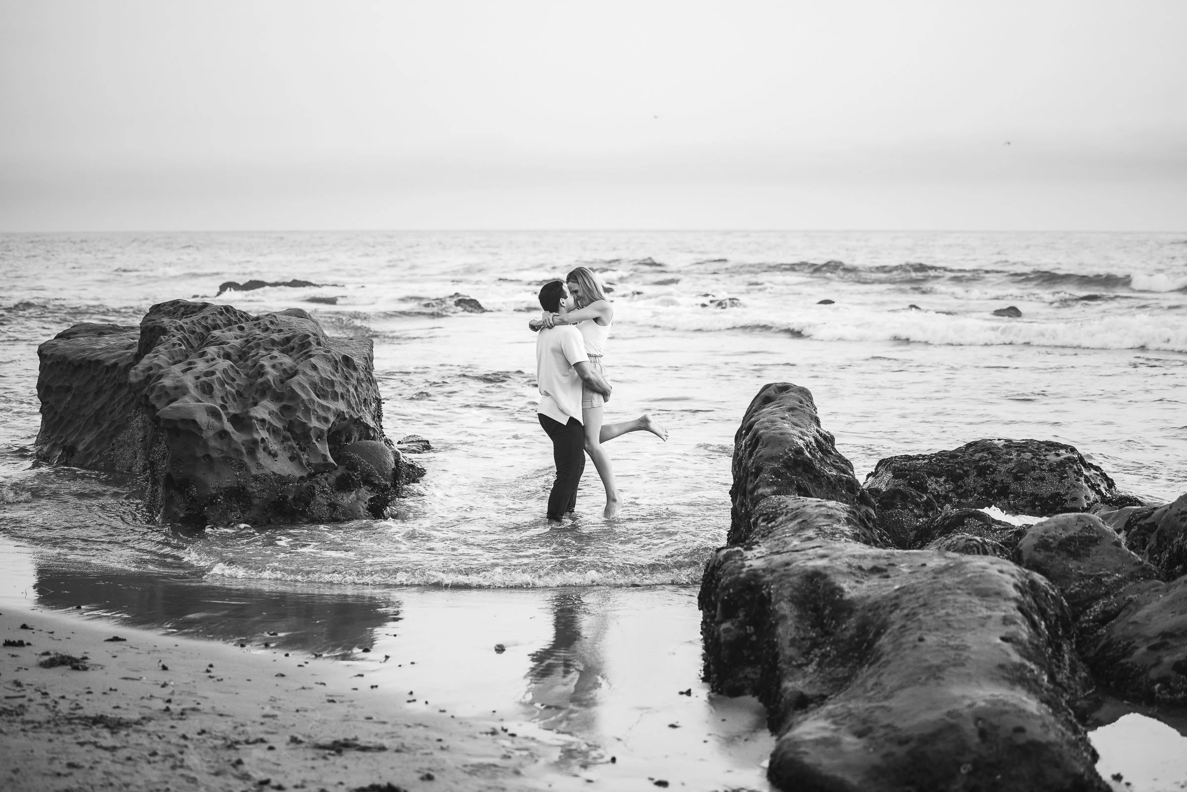 A couple is standing in shallow ocean water between large rocks on a beach. The man is holding the woman, who has her arms around his neck, and she is lifting her leg while they look at each other.