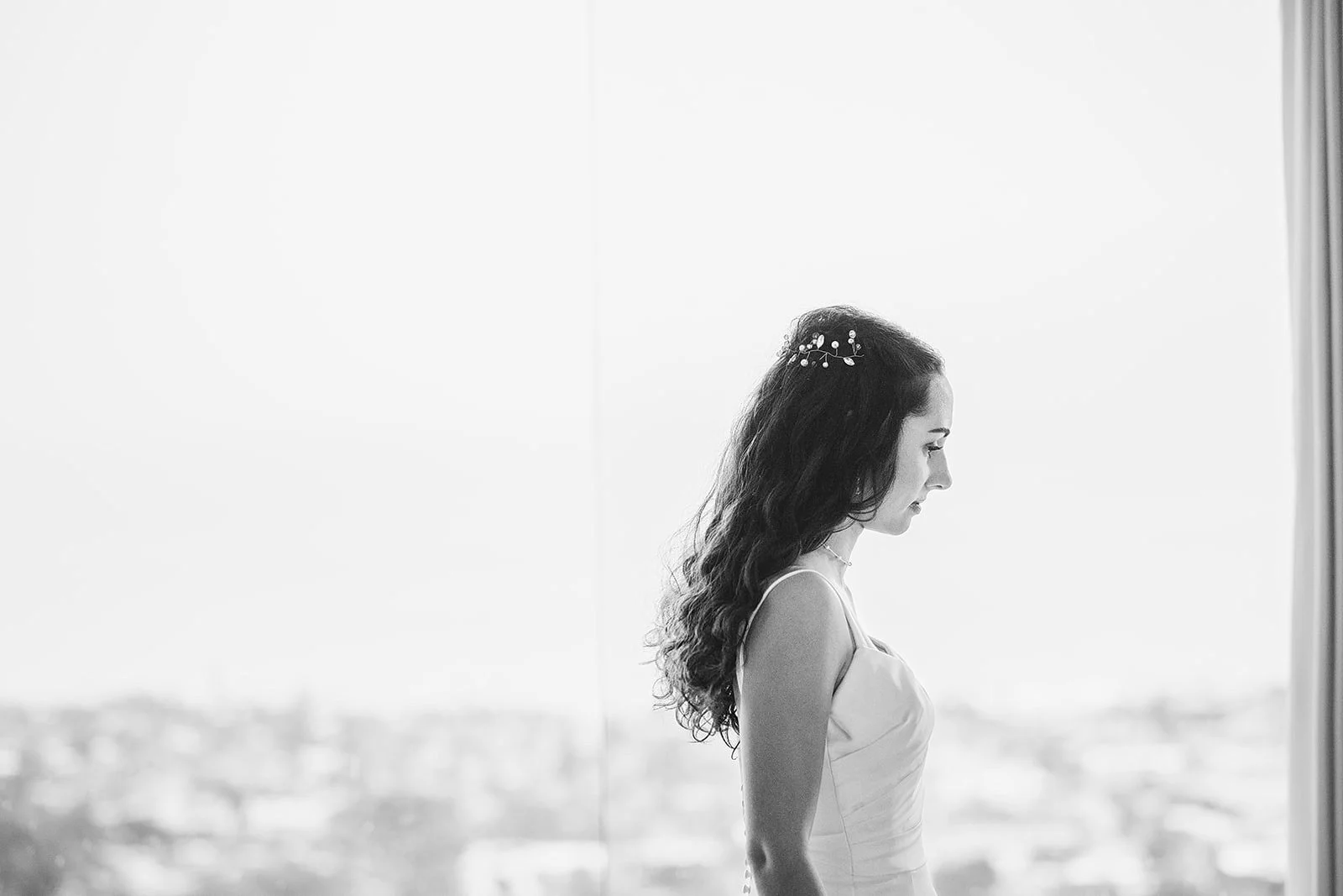 Black and white photo of a woman with long curly hair wearing a dress, standing indoors near a large window with a cityscape view outside, facing sideways.