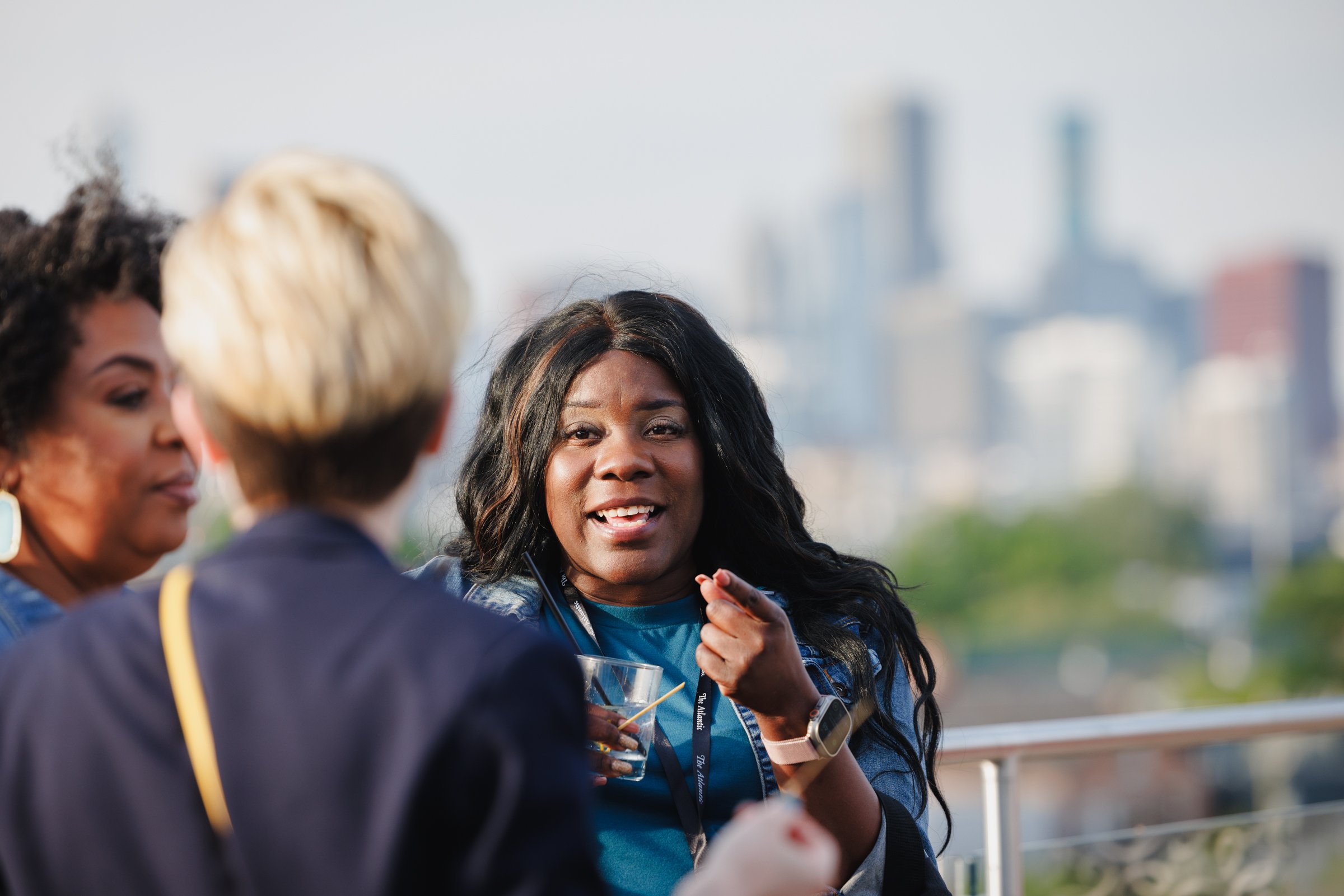 Three women engaged in conversation on a rooftop with a city skyline in the background. One woman in the center, with long black hair, is speaking and holding a drink, while the other two women listen.