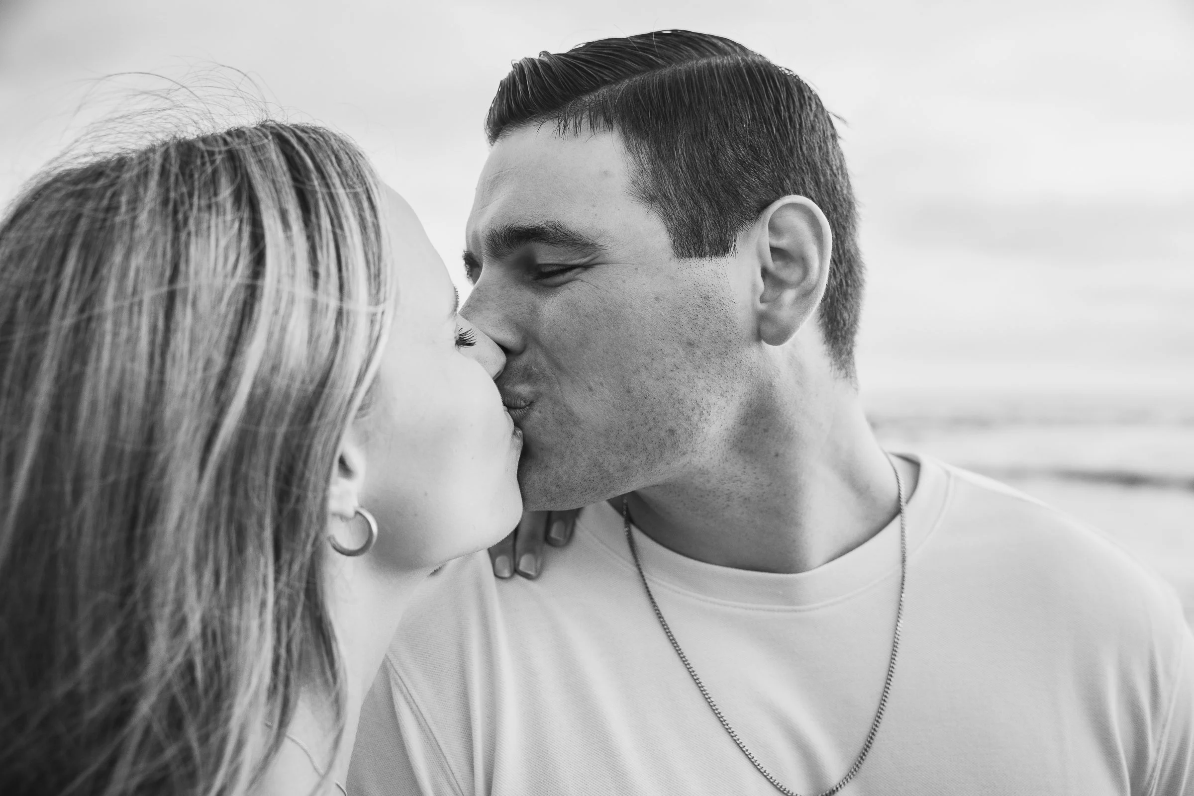 A black-and-white photo of a couple kissing near the beach, with the ocean in the background.