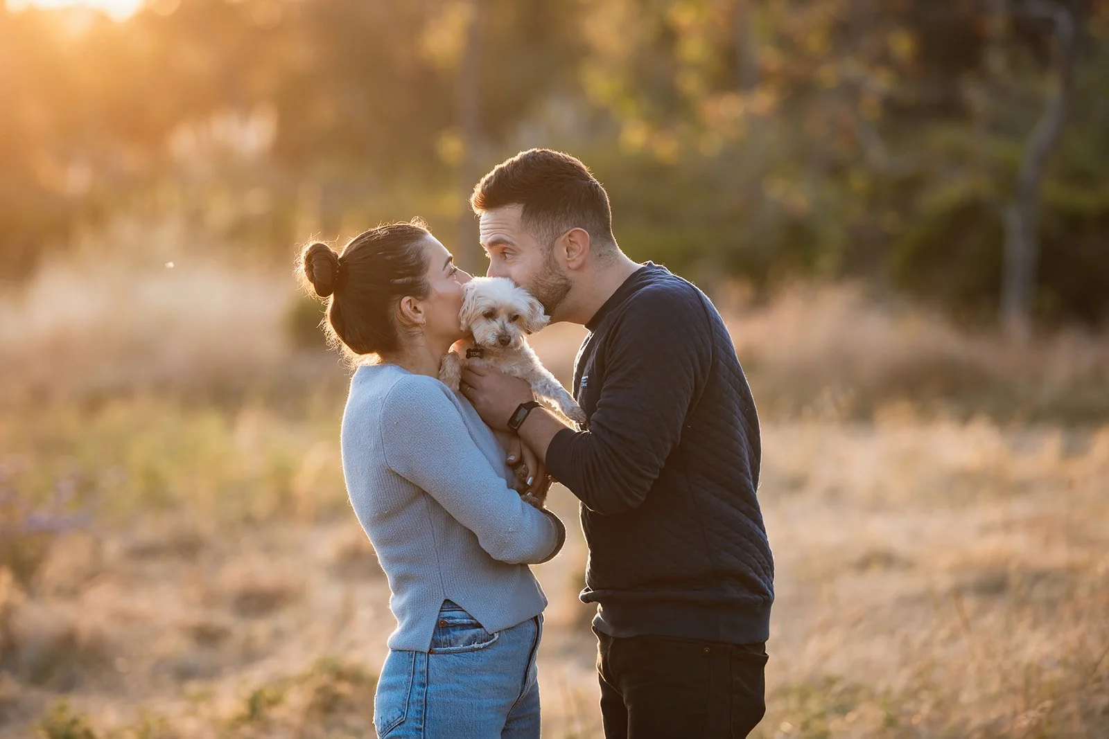 A couple and their small white dog kissing outdoors during sunset, in a field with trees in the background.