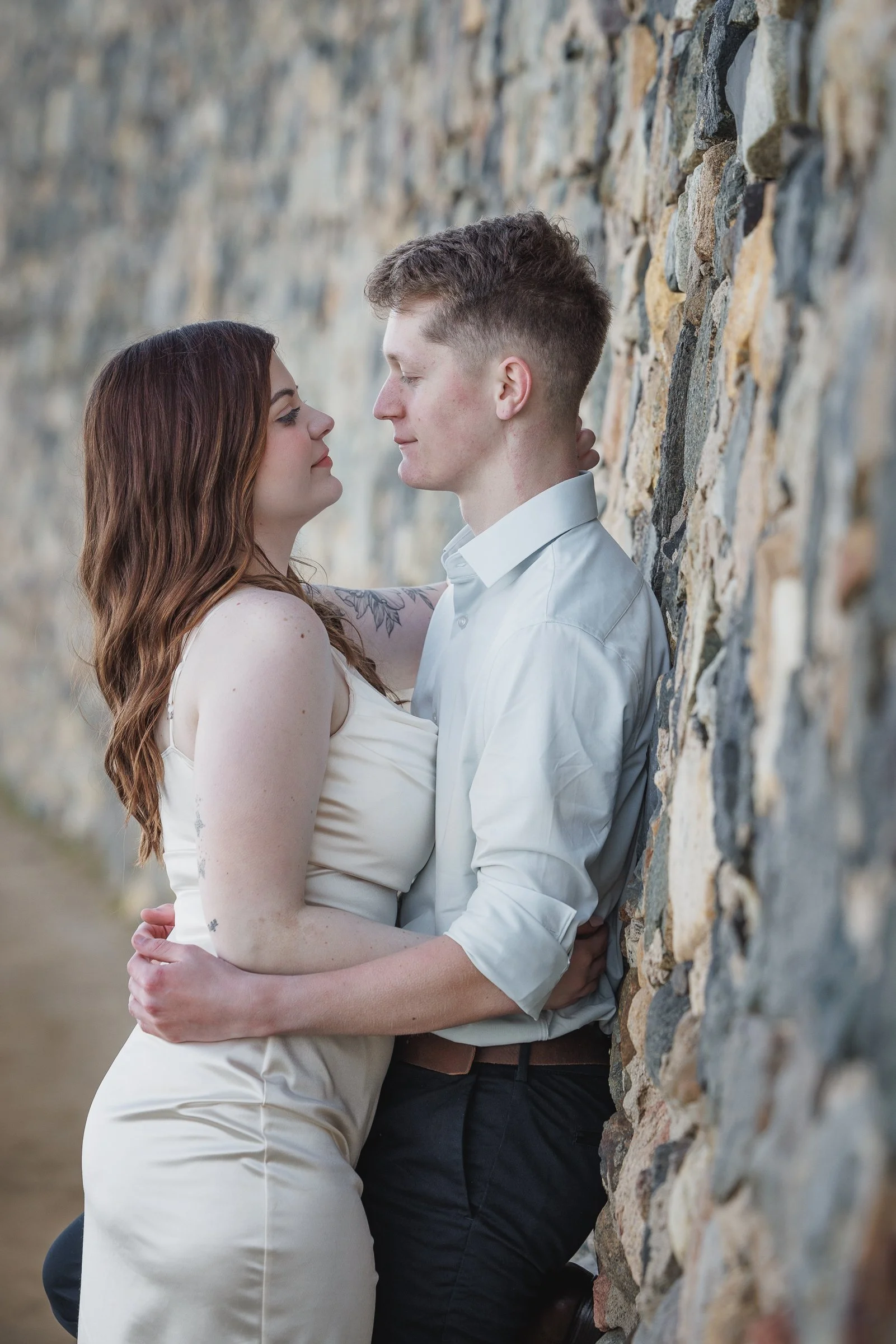 A couple stands close to each other against a stone wall, gazing into each other's eyes.