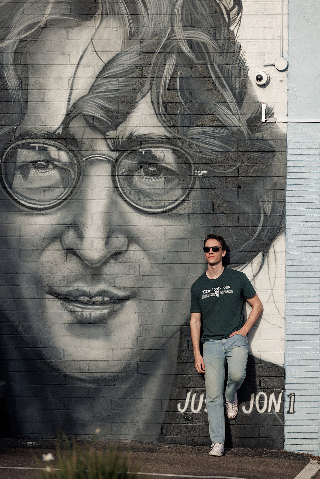 A young man is standing against a wall featuring a large black and white mural of a woman's face wearing glasses. The man is wearing sunglasses, a dark t-shirt, light jeans, and white shoes, and is leaning casually with one foot against the wall.