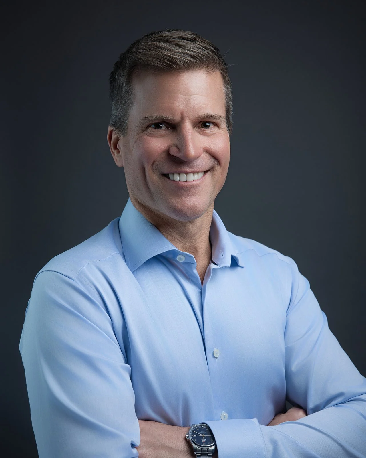 A portrait of a smiling man with short brown hair, wearing a light blue dress shirt and a silver watch, against a dark grey background.