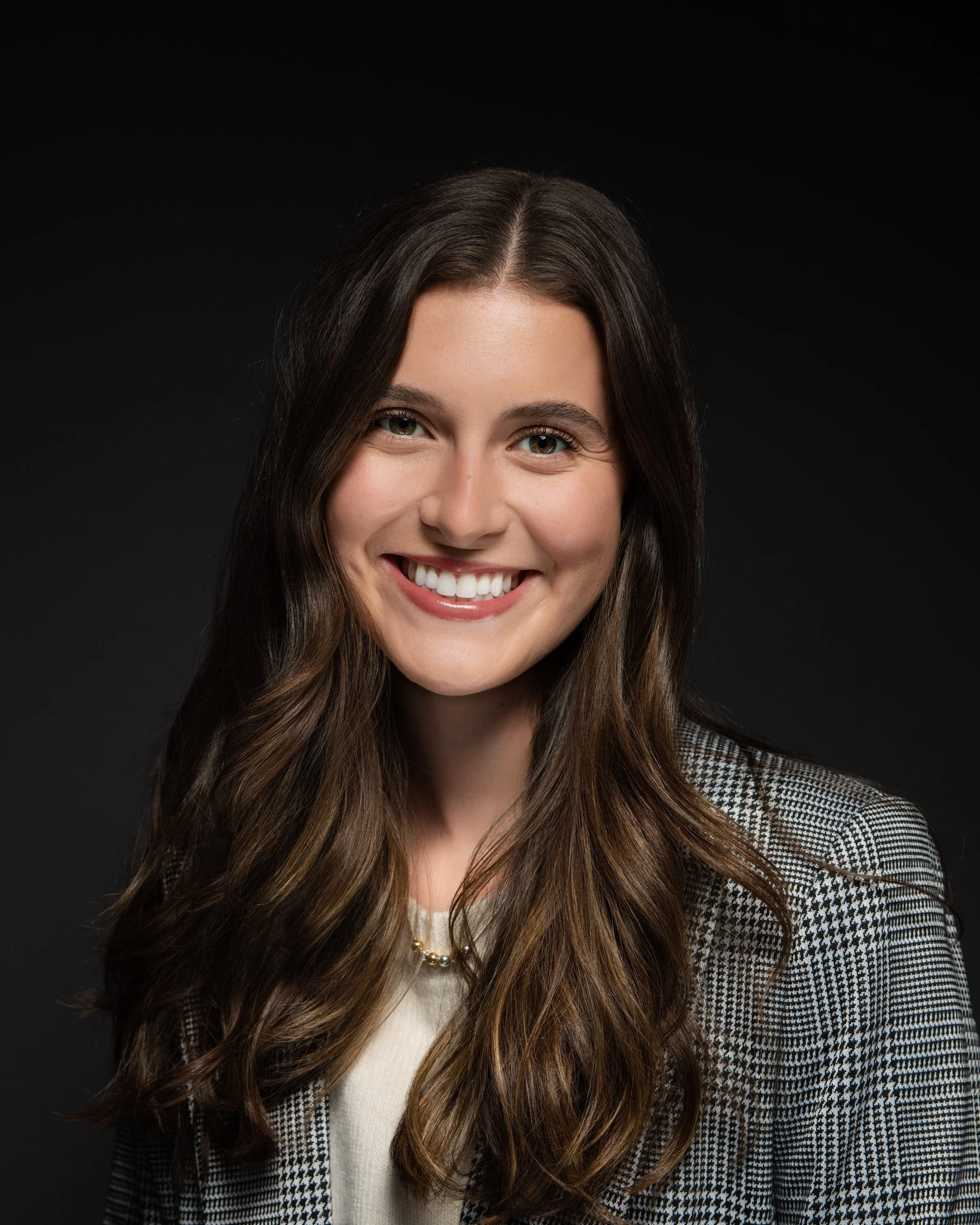 Smiling young woman with long brown hair, wearing a checkered blazer and a pearl necklace, against a black background.