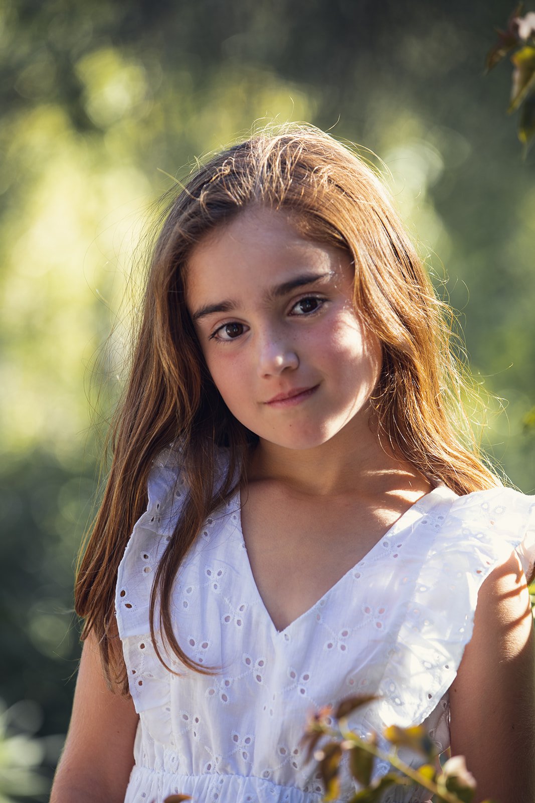 A young girl with long brown hair wearing a white dress with eyelet details outdoors, with a blurred green background and sunlight.