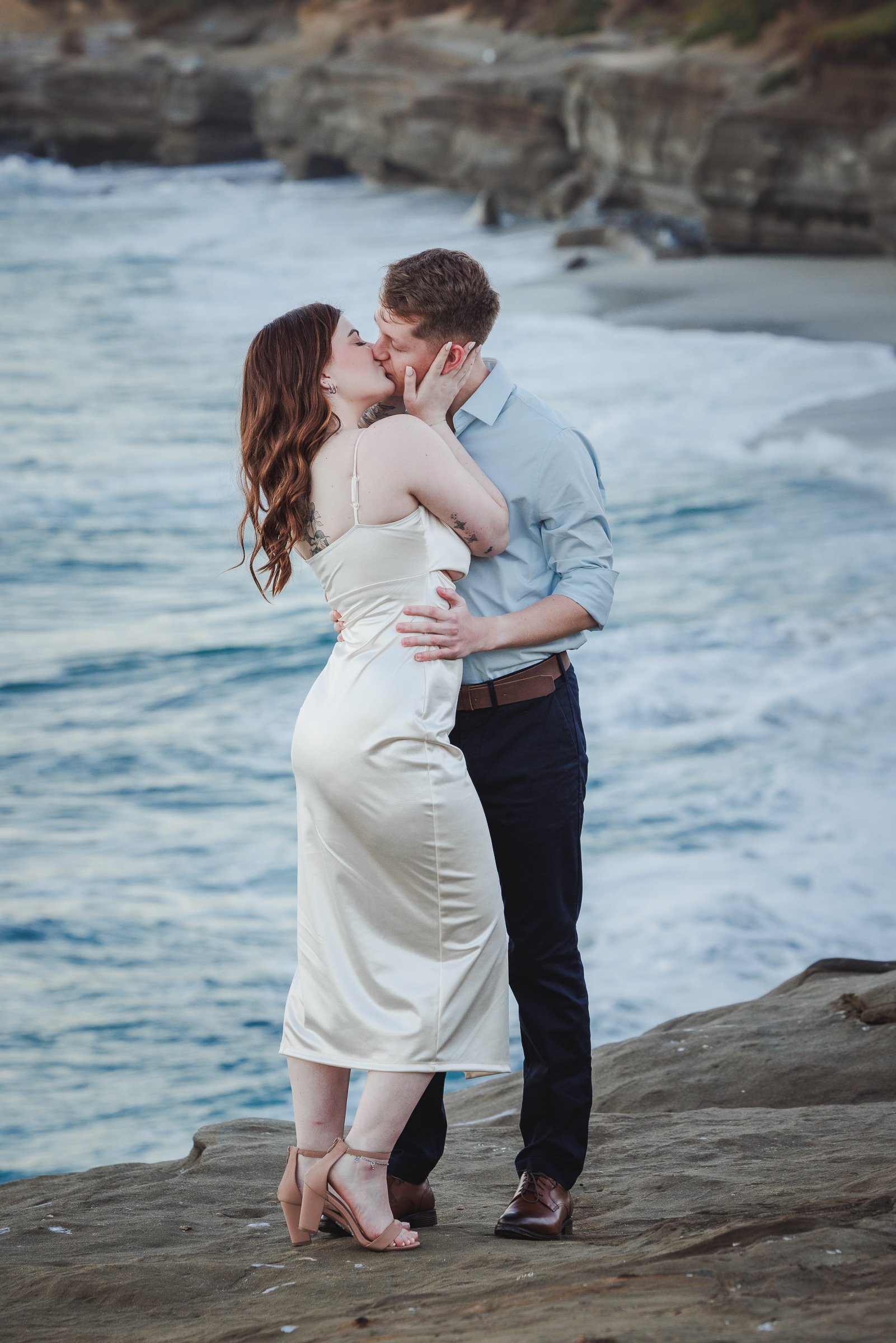 A couple kissing on a rocky shoreline with ocean waves and cliffs in the background.