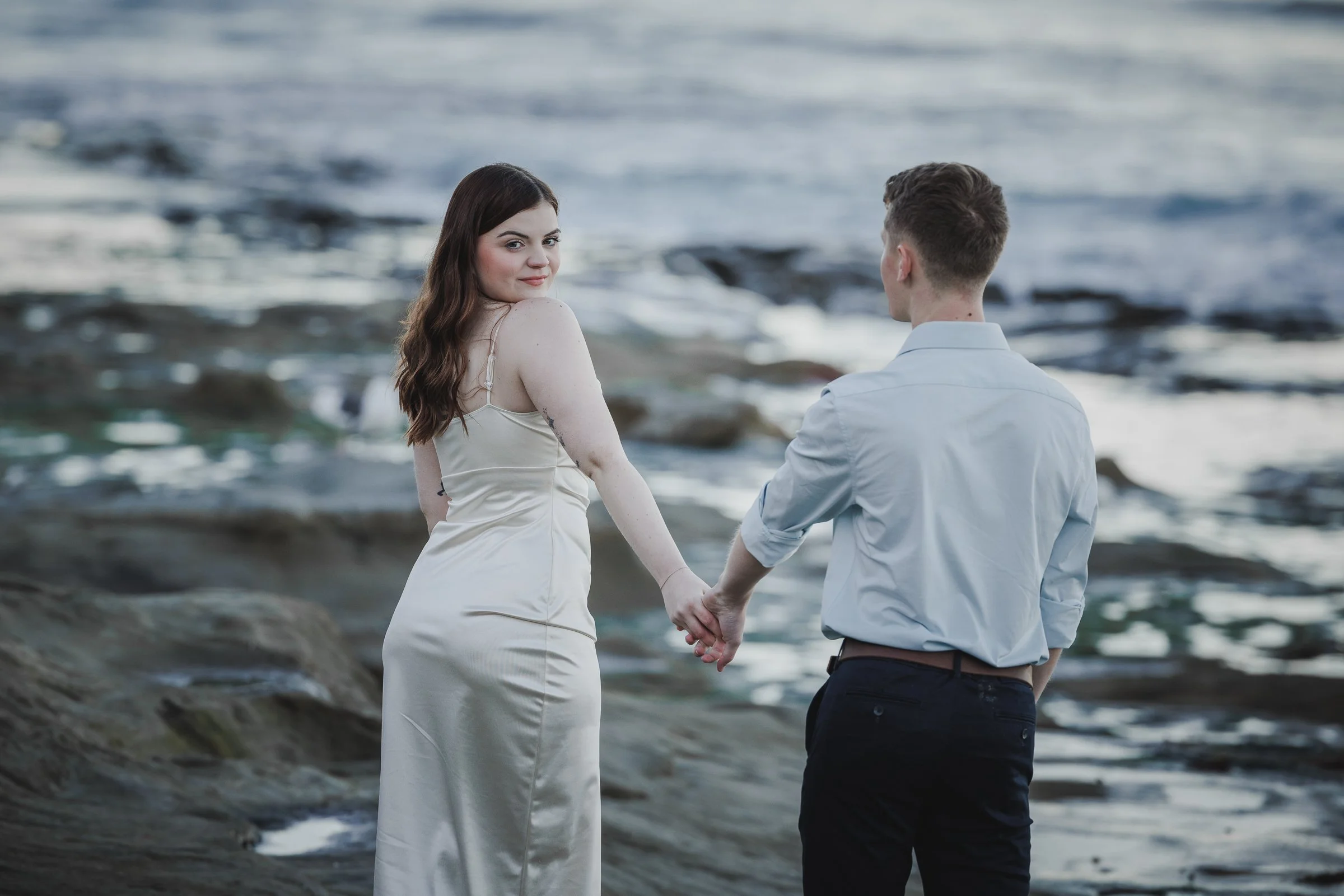 A young couple holding hands on a rocky beach with the ocean in the background. The woman is looking back and smiling at the camera, wearing a satin dress, and the man is facing away, dressed in a light-colored shirt and dark pants.
