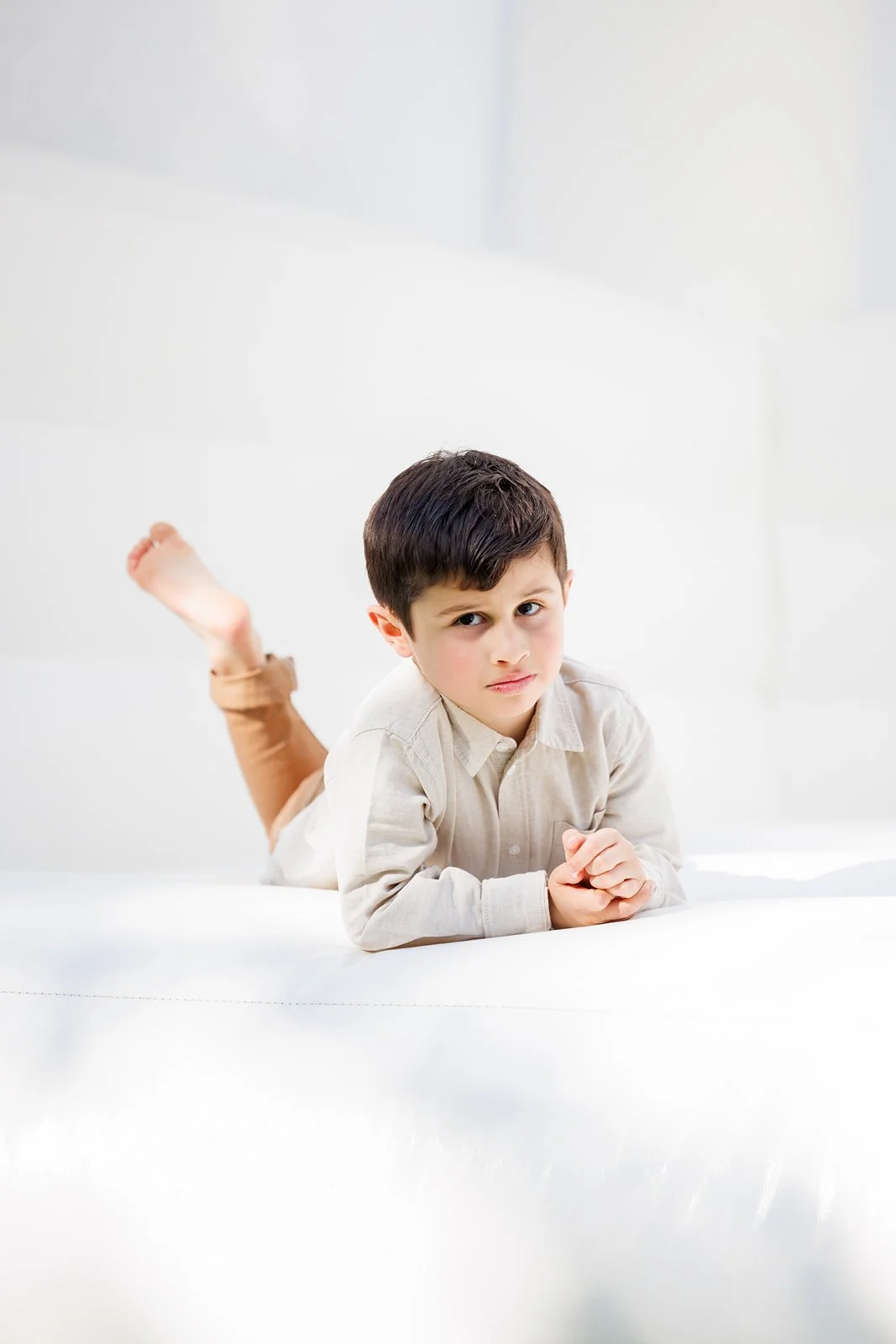 Child lying on stomach on a white surface, resting his chin on his hands, with a skeptical or confused expression.