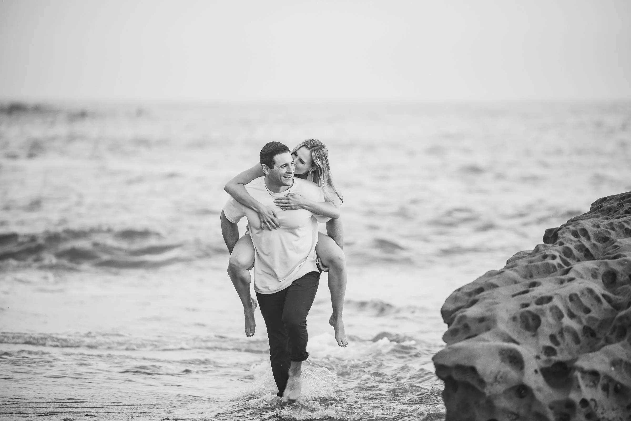 A happy couple at the beach, with the woman giving the man a piggyback ride as they walk through the shallow water near rocks.