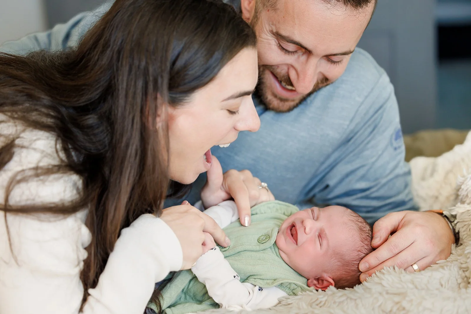 A woman and a man smiling and looking at a crying baby lying on a soft blanket, with the woman pointing at the baby.
