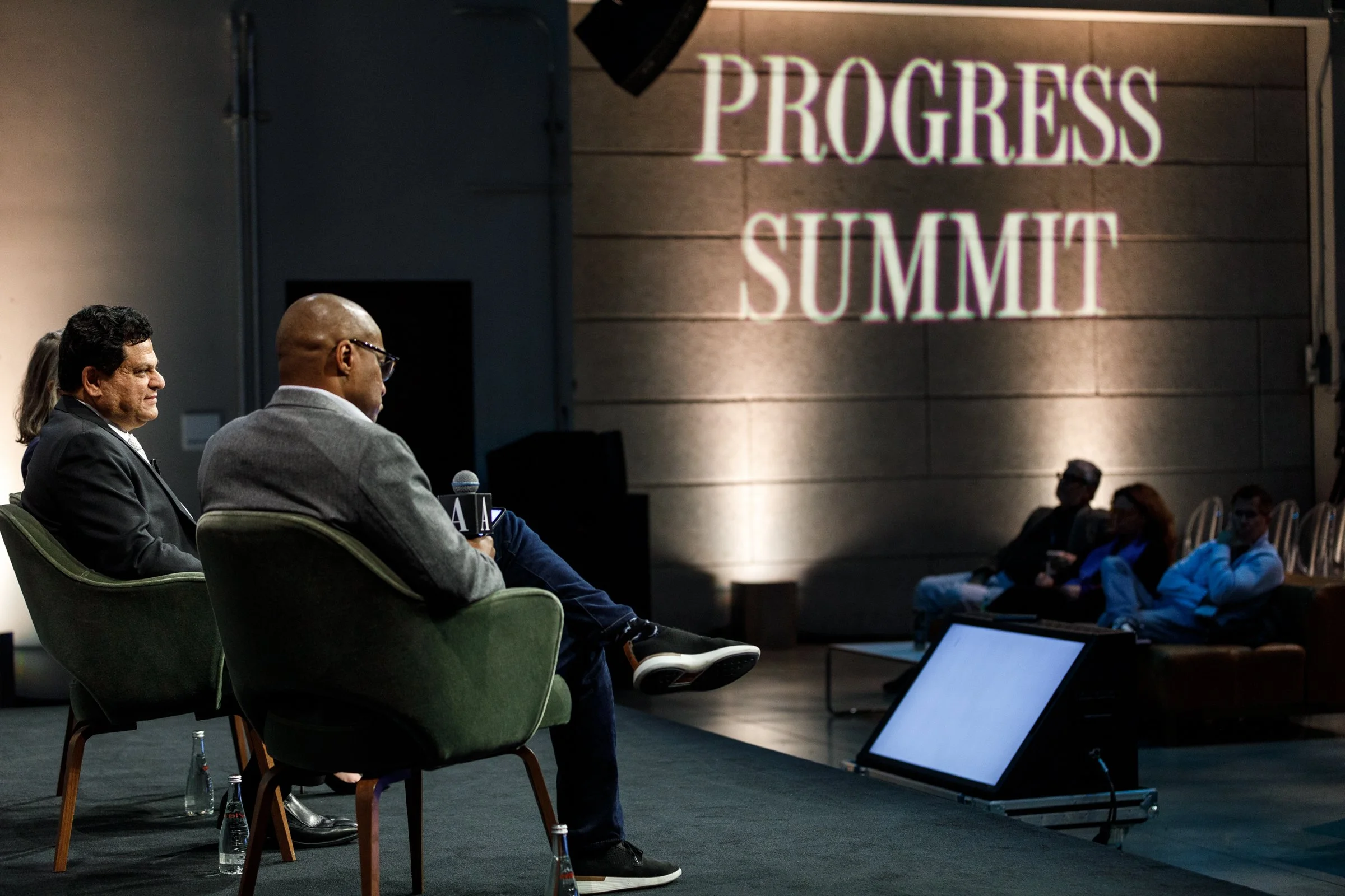 Panel discussion at a conference titled 'Progress Summit' with leather chairs on a stage, and a large screen on the floor in front.