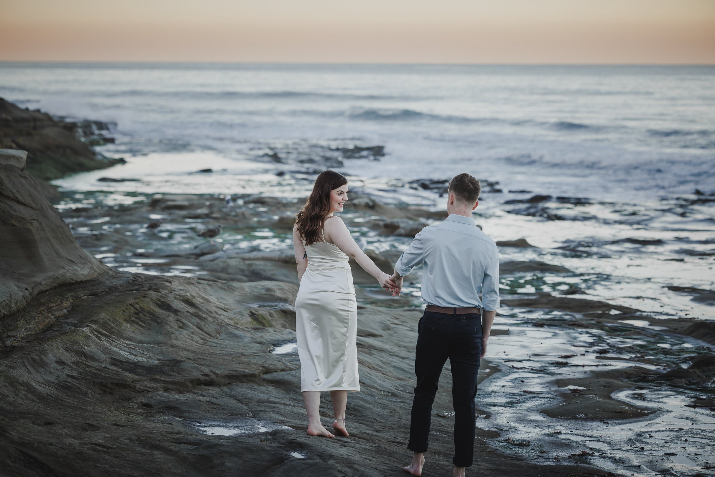 A couple holding hands on a rocky beach during sunset, with the woman dressed in a white dress and the man in a light blue shirt and black pants.