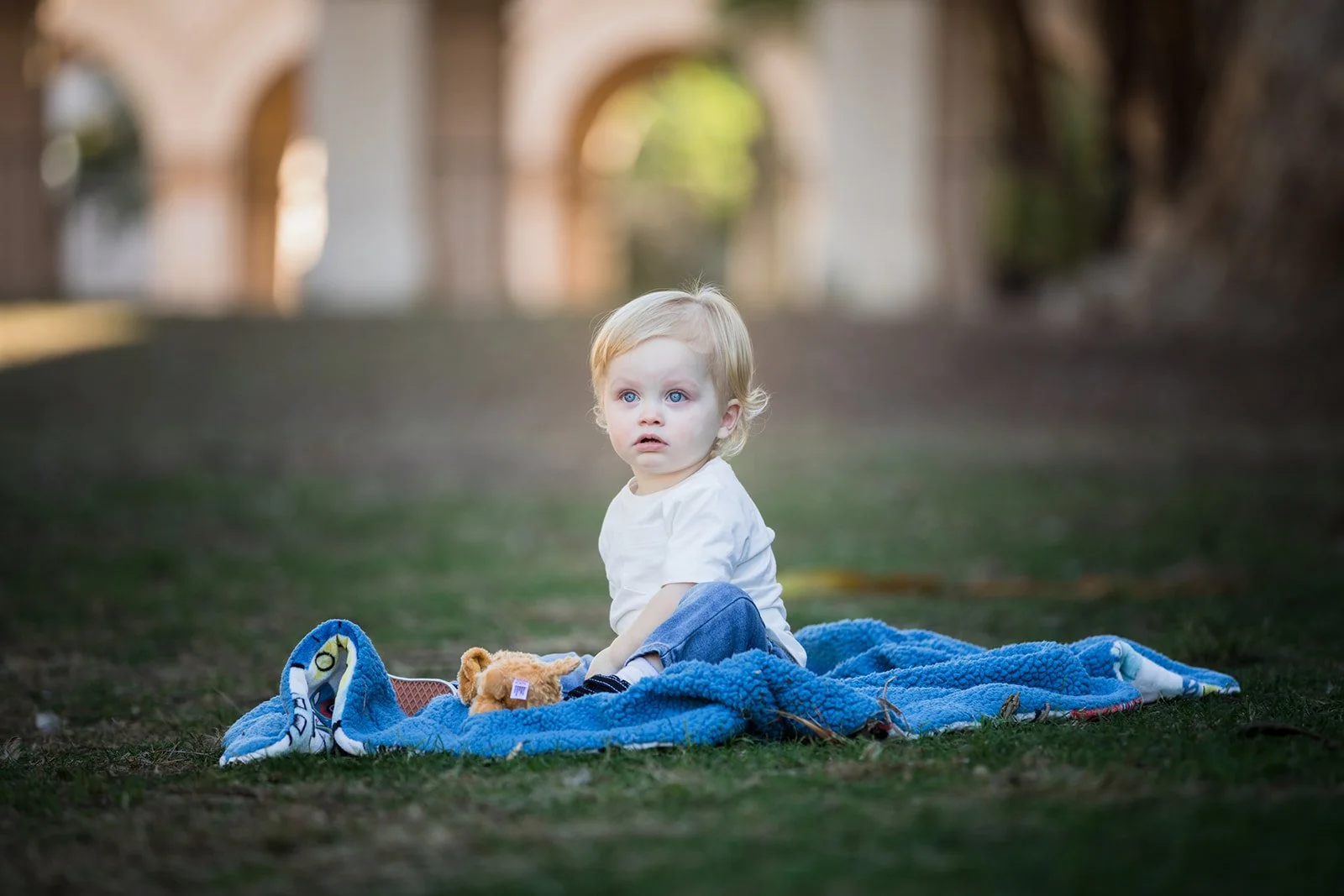 A young child with blonde hair and blue eyes sitting on a blue blanket on the grass in a park, with a blurred background of trees and arches.