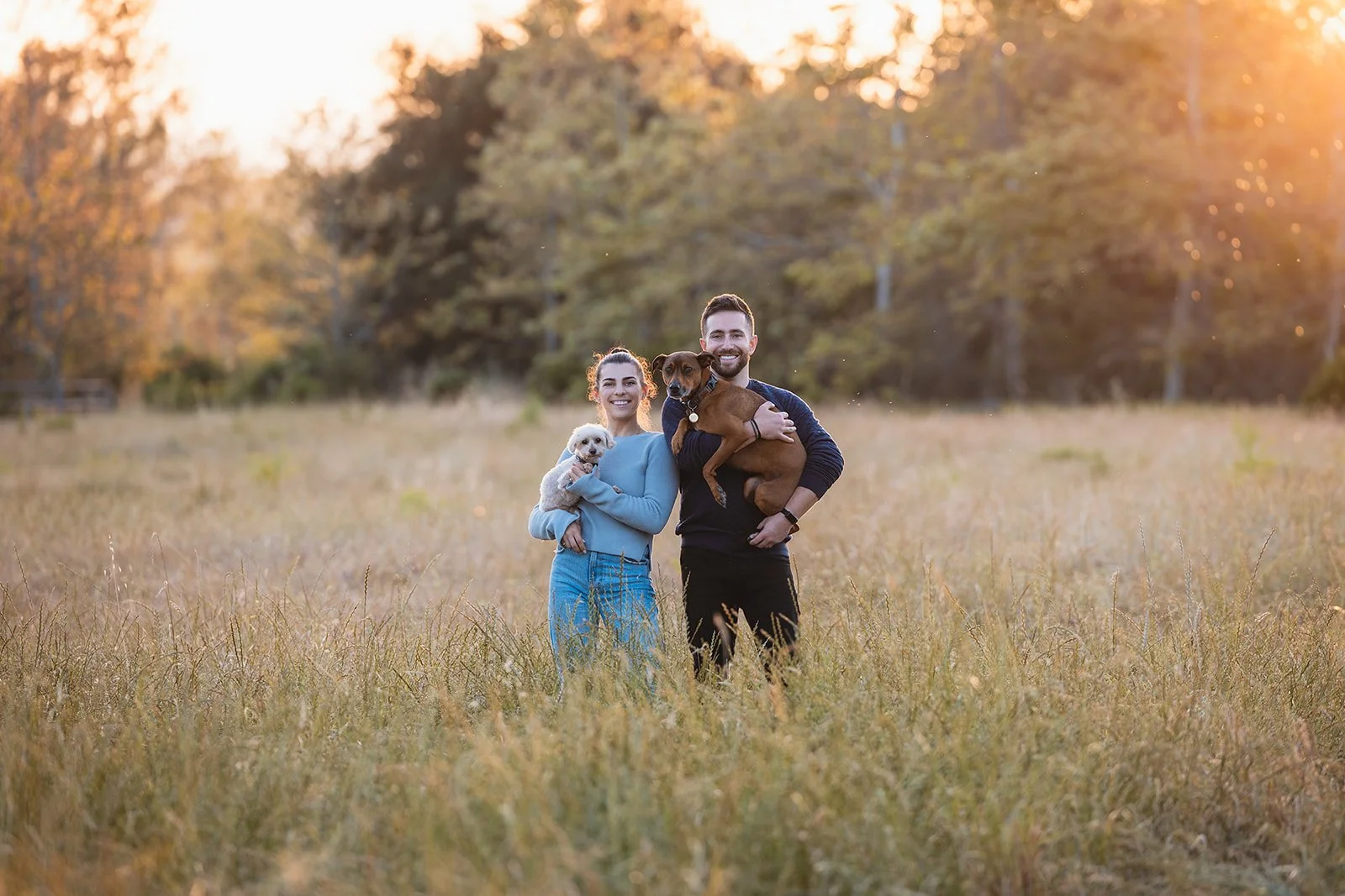 A happy couple with two dogs standing in a grassy field during sunset.