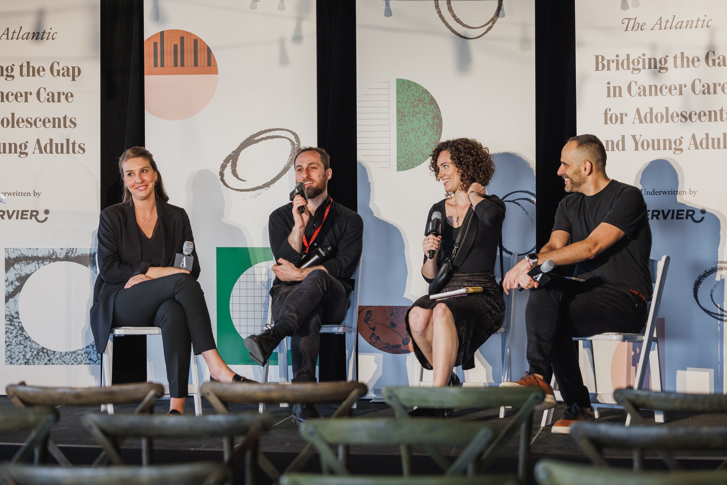 Four people sitting on a stage, engaged in a panel discussion at a conference, holding microphones. The background features graphics and text about cancer care for adolescents and young adults.