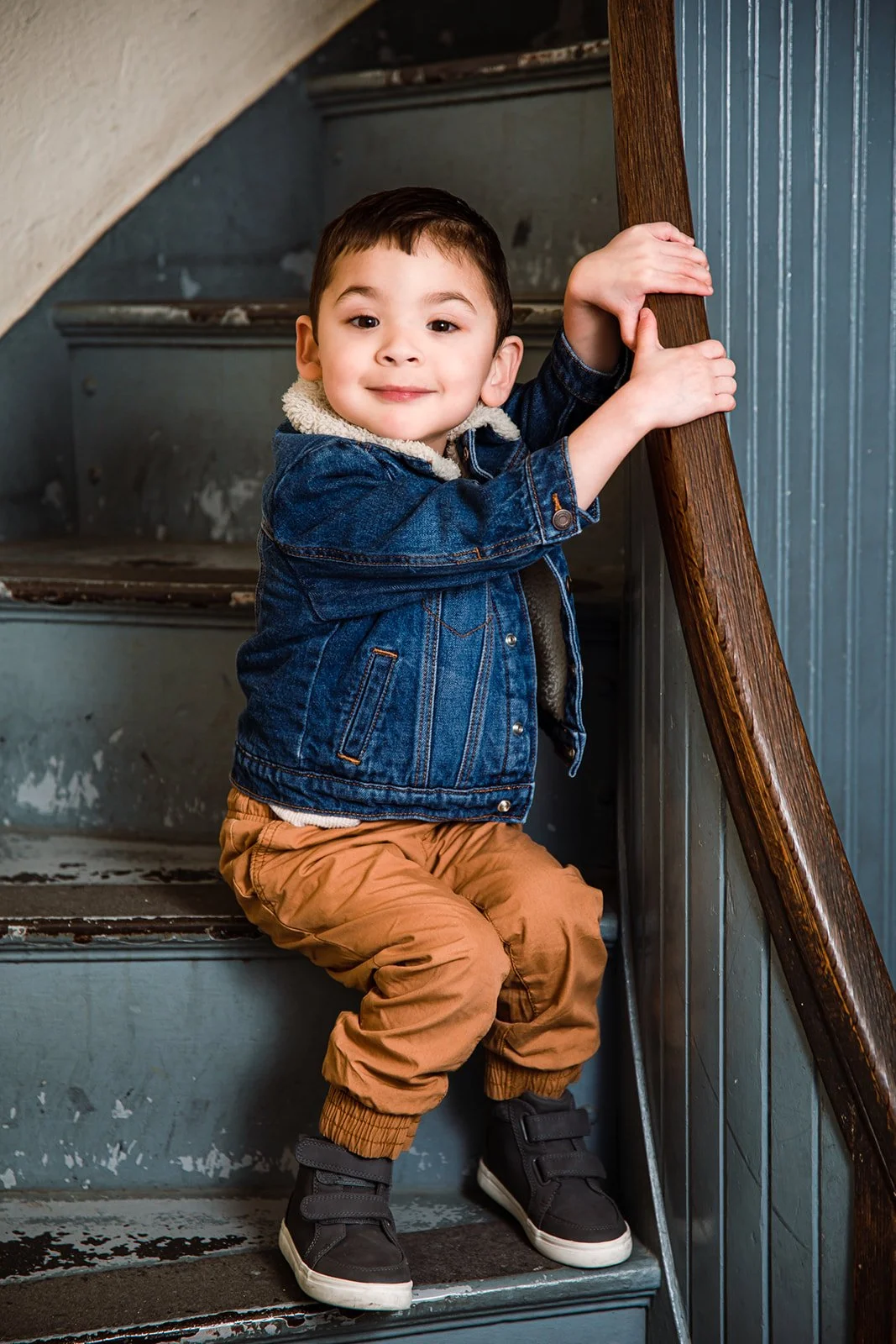 A young boy sitting on a worn staircase, holding the wooden railing and smiling at the camera. He is wearing a denim jacket, tan pants, and black sneakers.