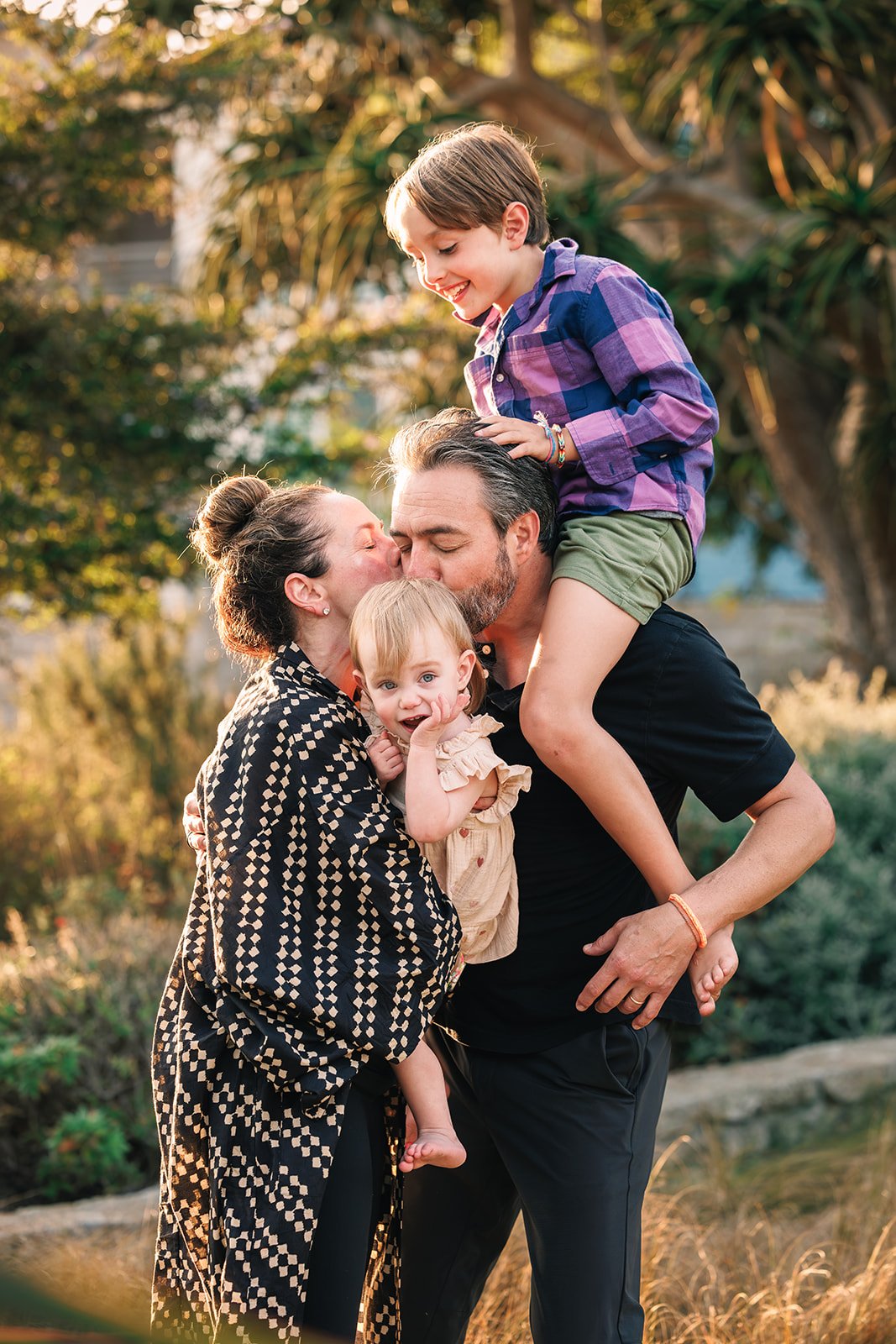 A family of four enjoying an outdoor moment, with a woman kissing a man on the cheek while a young boy sits on the man's shoulders, and a small girl holding onto the man's arm, all smiling and surrounded by trees and warm sunlight.