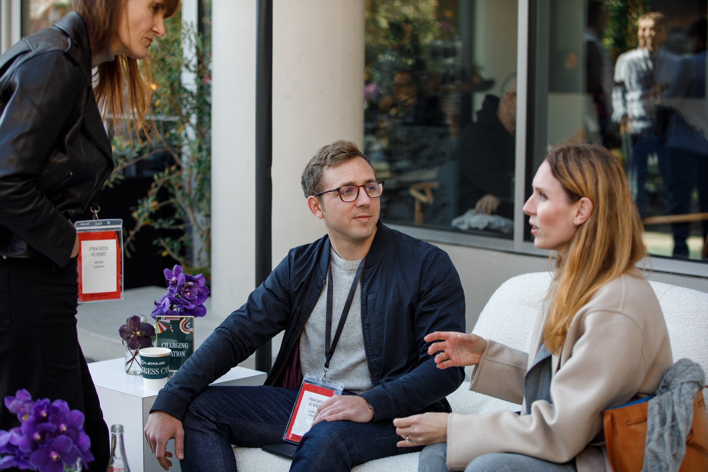 Two people sitting and talking on a white couch in an outdoor area during daytime, with a woman standing and speaking to them. They have conference badges around their necks.