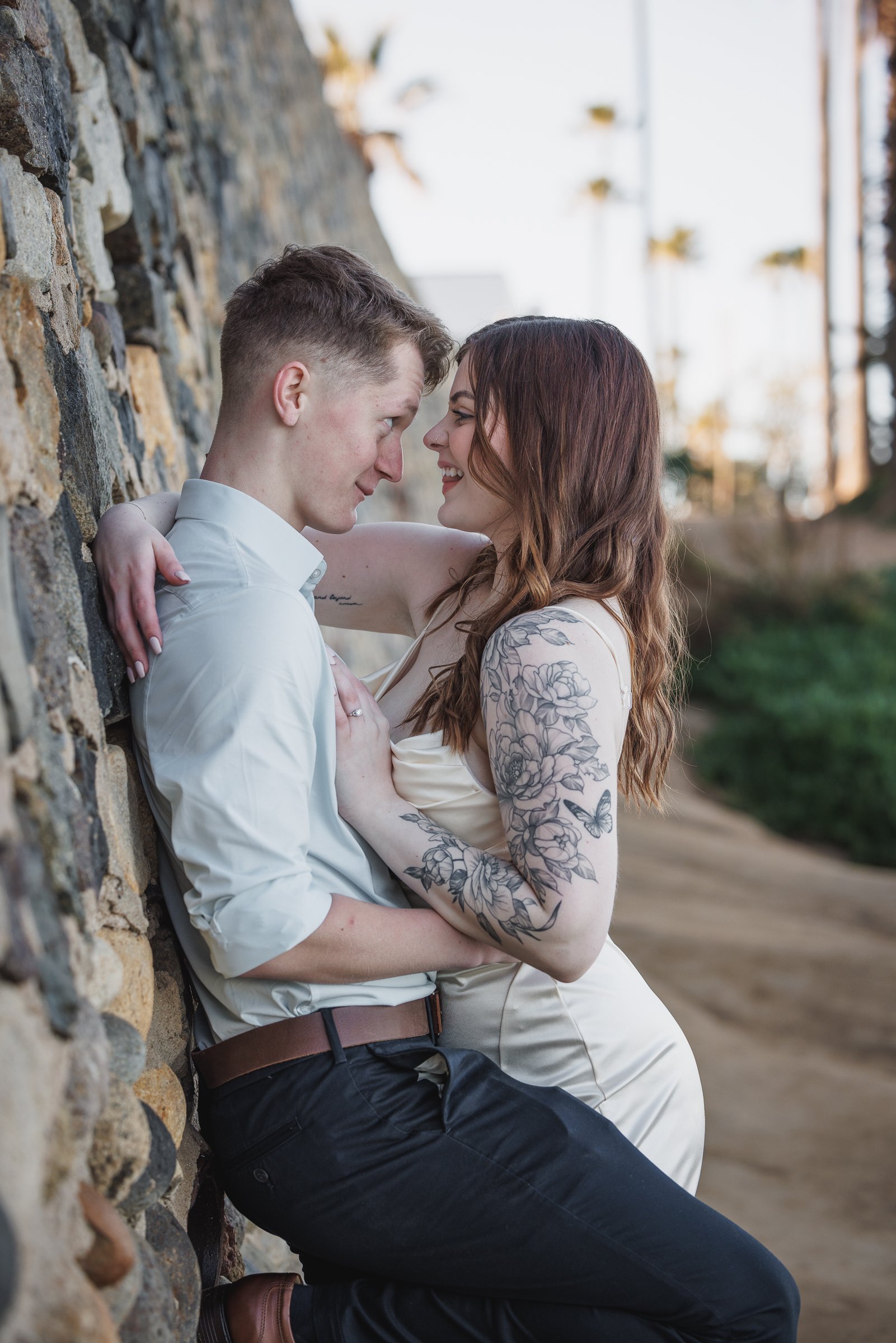 A couple leans against a stone wall, close with foreheads touching, smiling at each other outdoors during daytime.