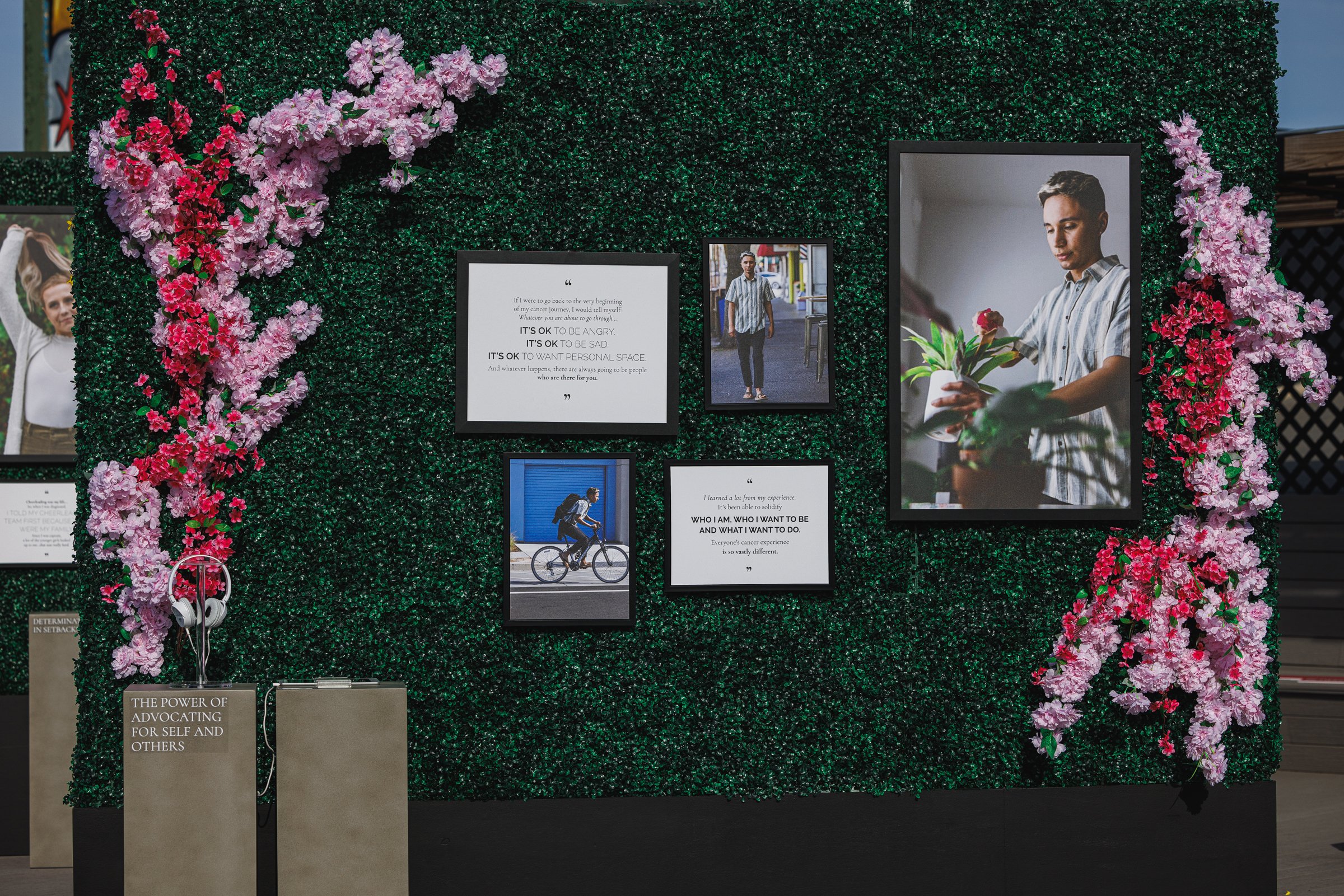 A green wall decorated with pink and purple flowers, featuring framed photos and quotes. There are several framed pictures of a young man, one of him walking, one of him holding a flower, and a bicycle rider. A headphone stand with a sign about self-