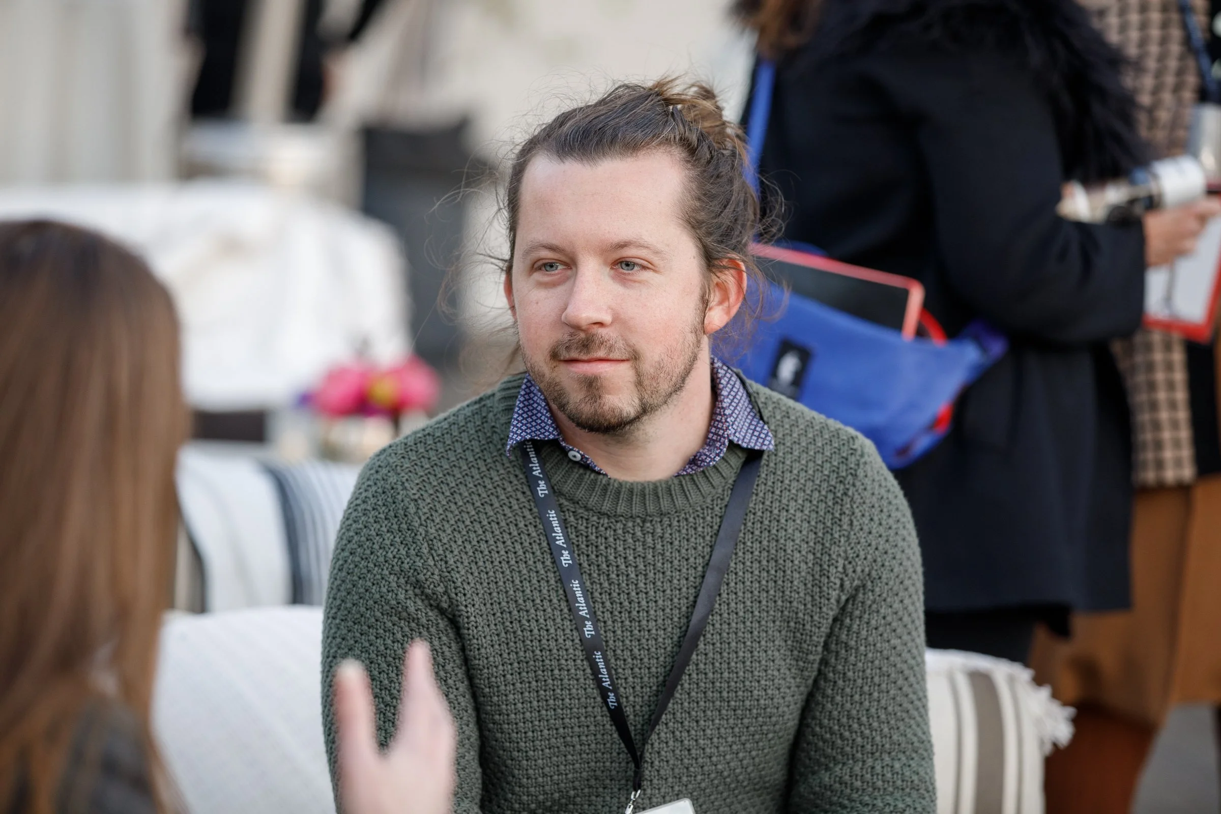 A man with long hair, beard, and mustache wearing a green sweater and a lanyard with a badge, engaged in conversation at an outdoor event.